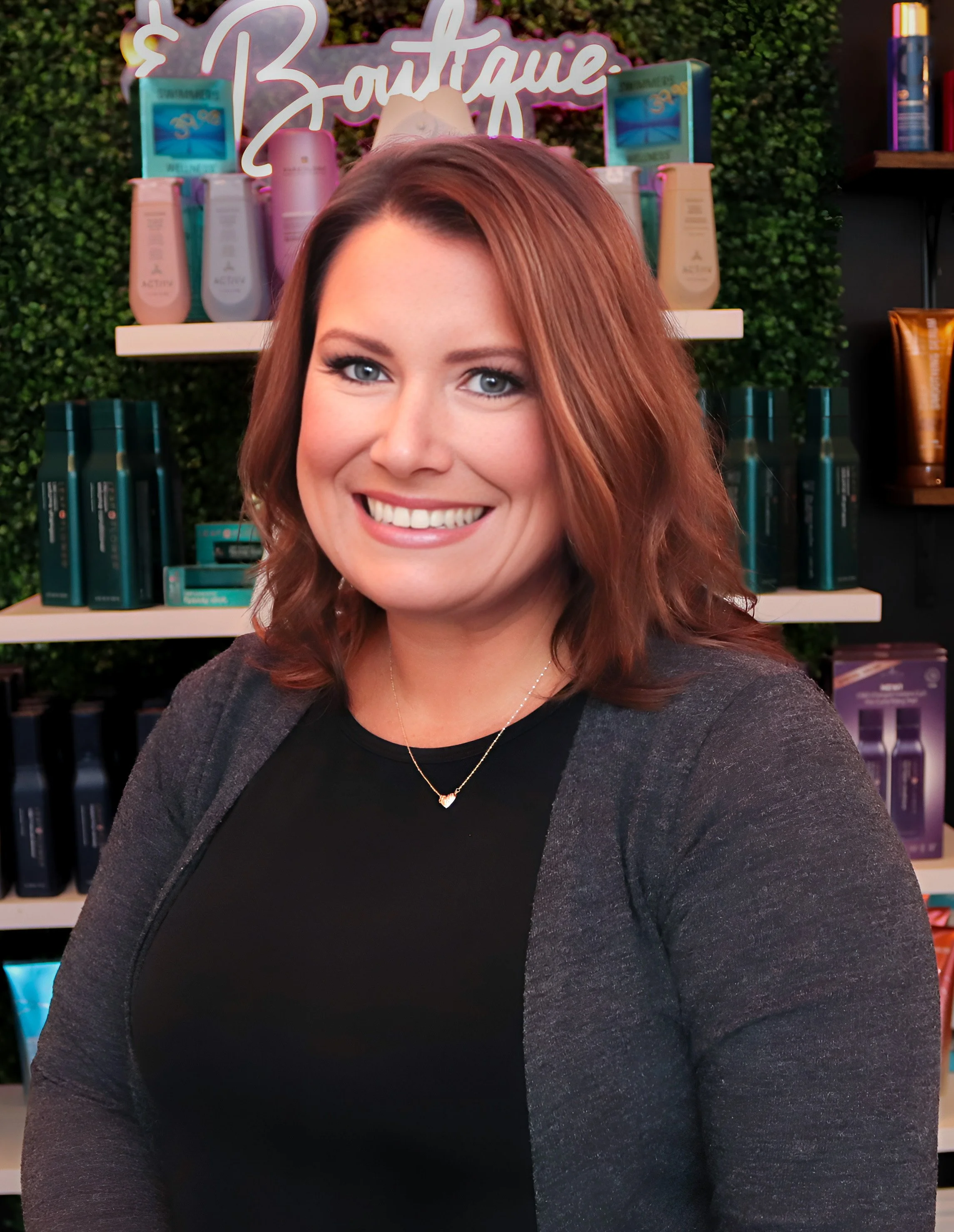 A woman with shoulder-length wavy hair smiling in front of shelves with hair care products in a boutique store.