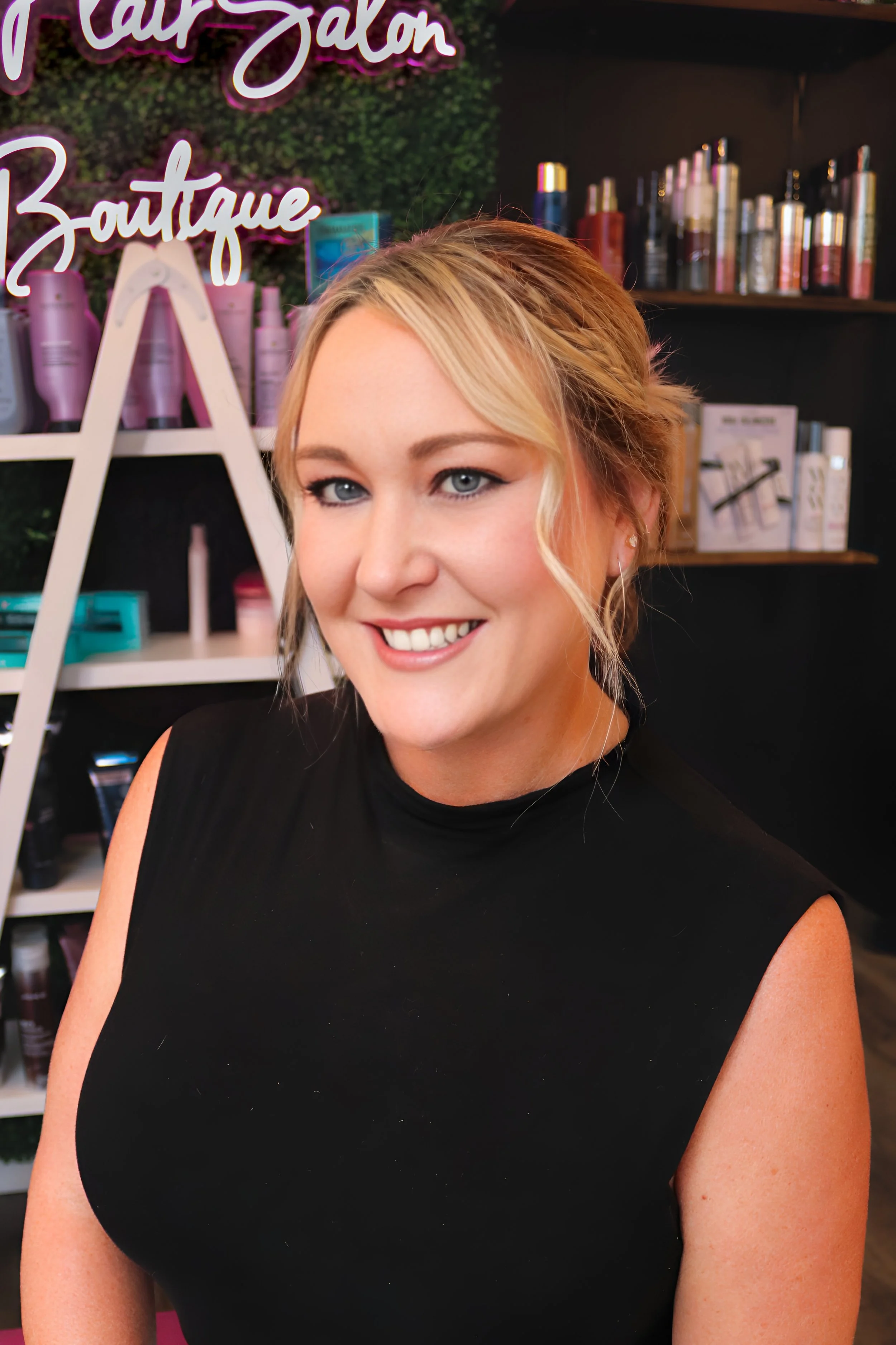 A woman smiling with her hair styled in an updo, wearing a sleeveless black top, standing inside a salon with shelves of hair products in the background.