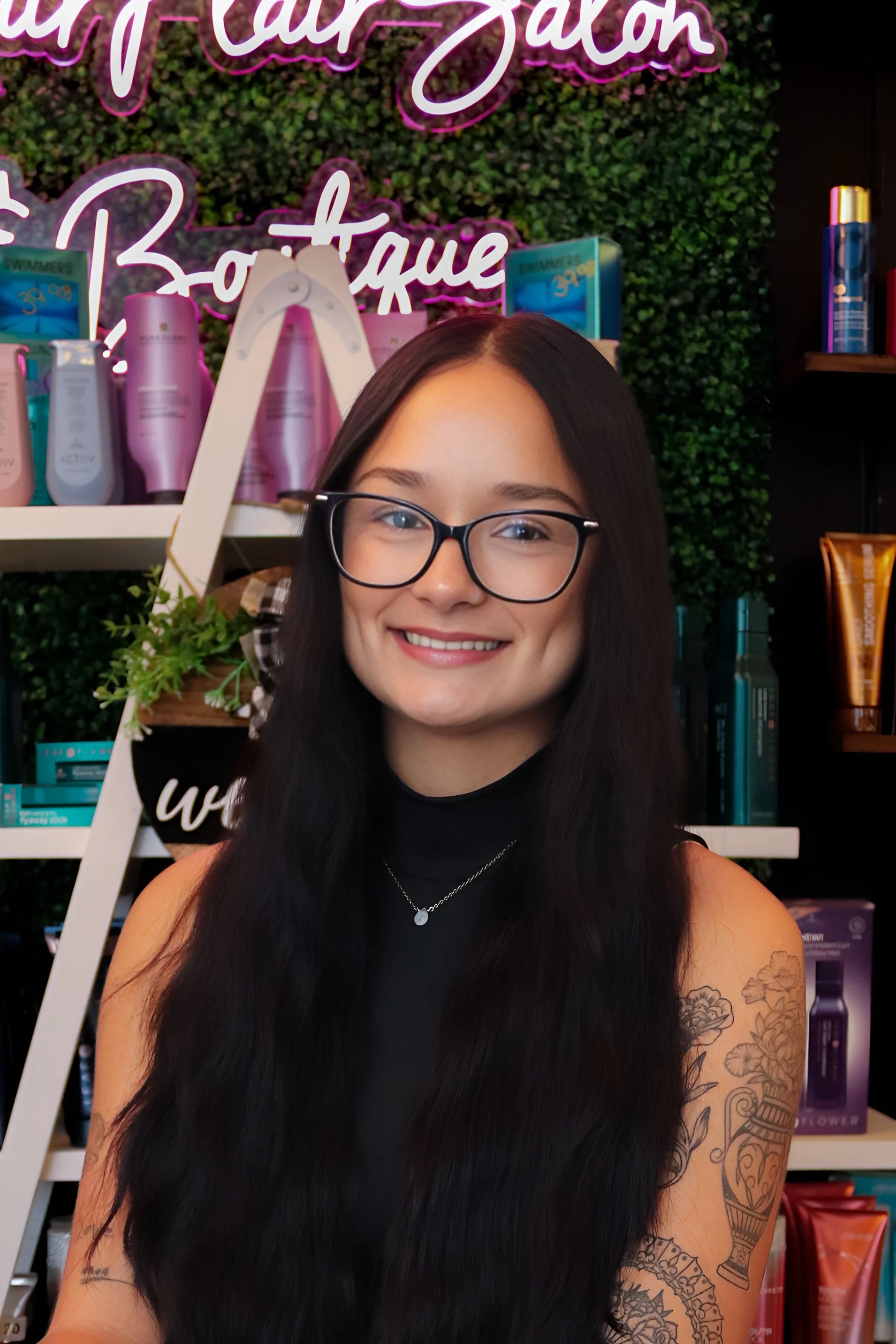 A young woman with long dark hair, glasses, and tattoos on her arms, smiling and posing in front of shelves of skincare or beauty products.
