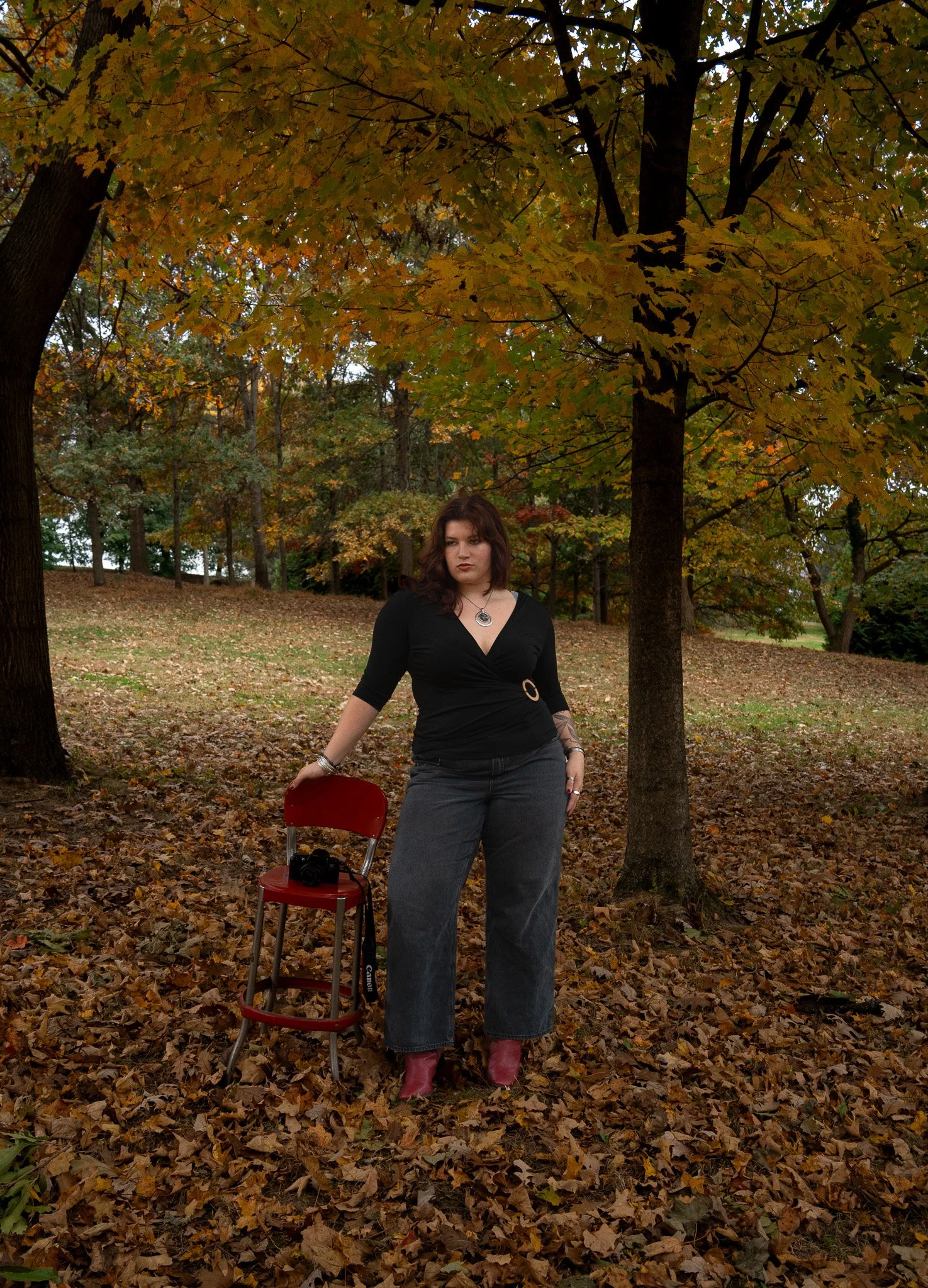 A woman standing outdoors in a park during autumn, surrounded by fallen leaves and trees with orange and yellow foliage, posing with one hand on a red chair that has a camera on it.