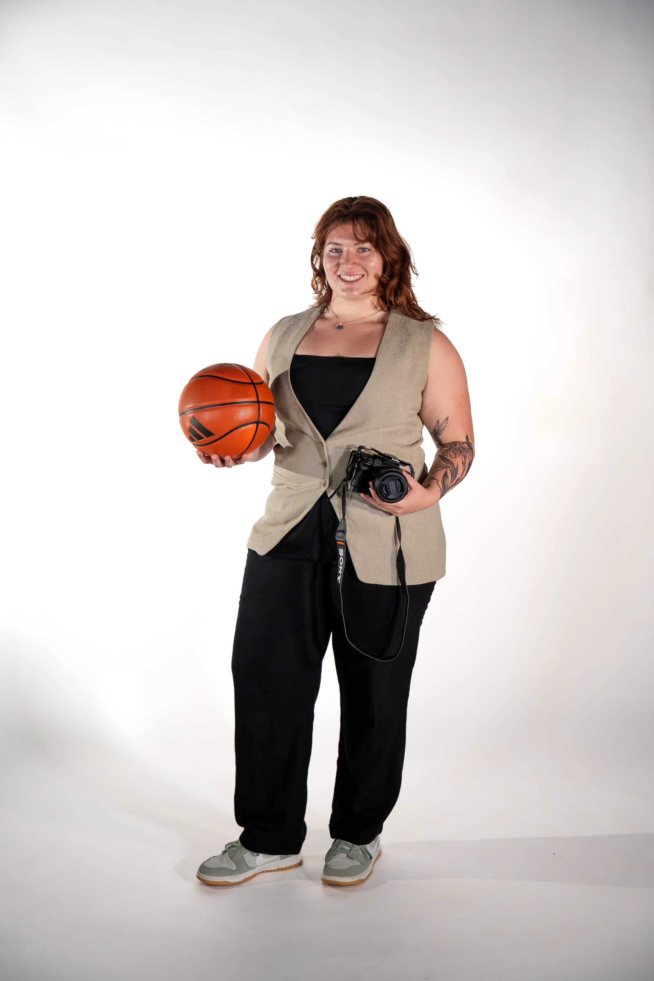 A woman with brown hair holding a basketball and a camera, wearing a sleeveless beige vest, black pants, and gray sneakers, standing against a white background.