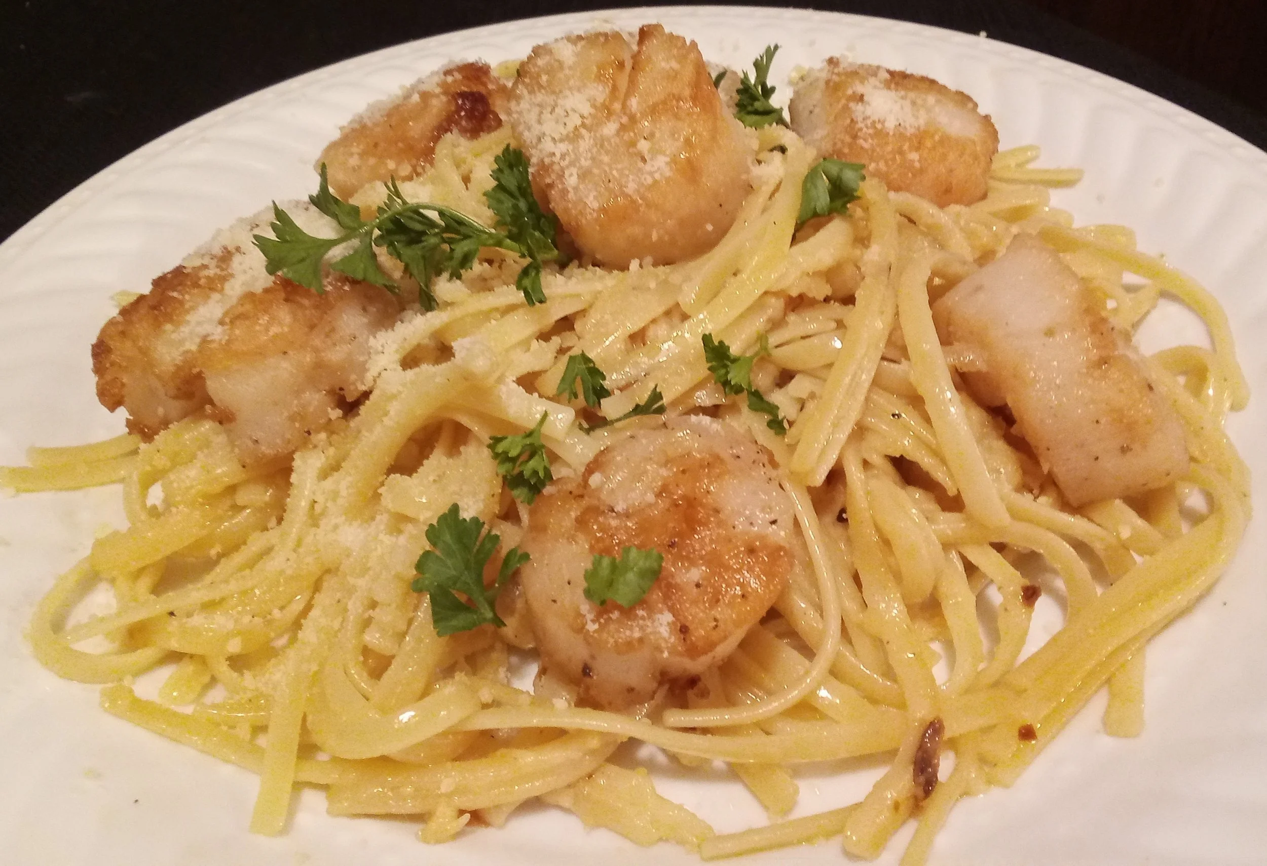 Spaghetti with fried scallops, topped with grated cheese and garnished with fresh parsley on a white plate.