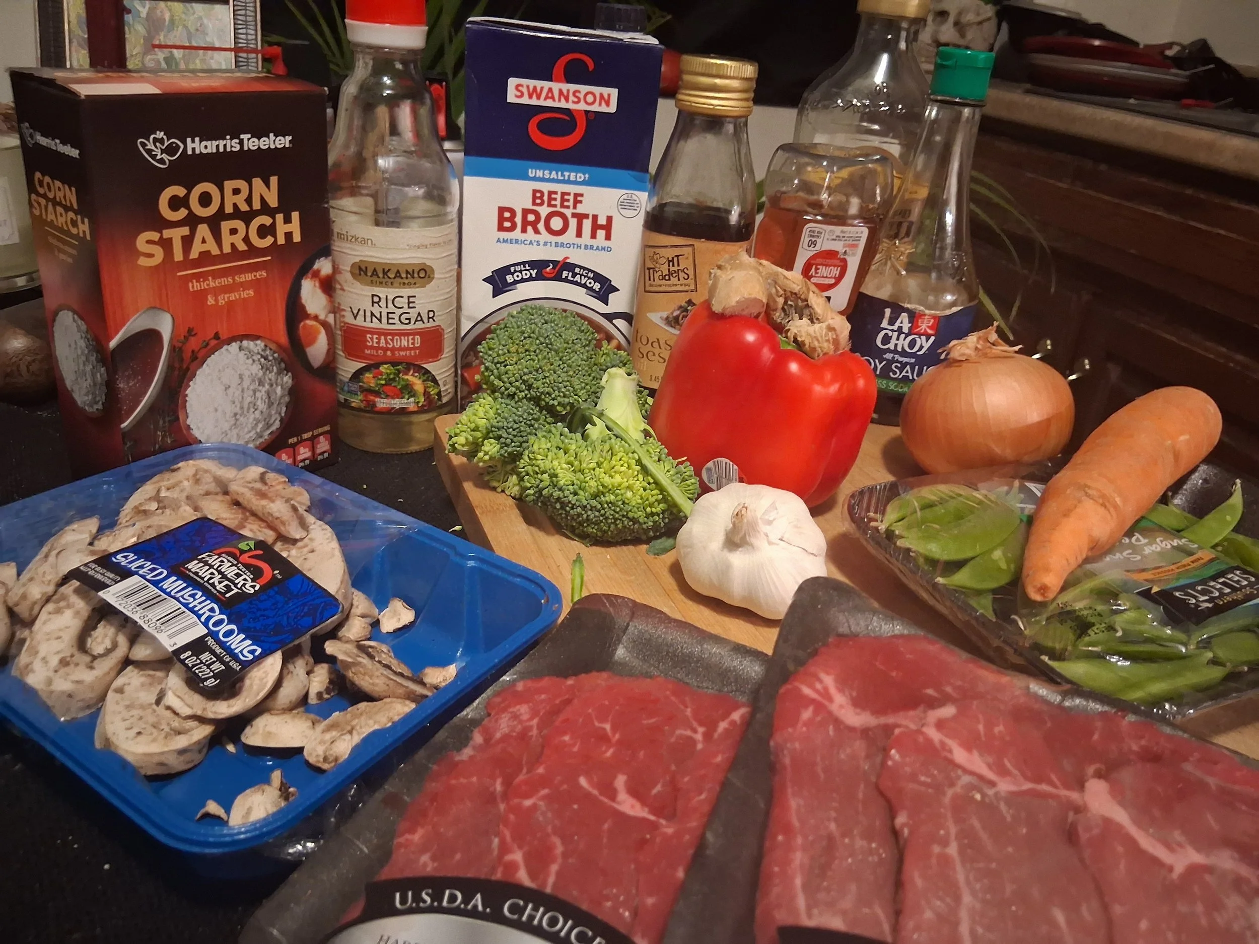 A variety of fresh and packaged ingredients for cooking, including beef slices, broccoli, red bell pepper, onion, garlic, carrot, sugar snap peas, sliced mushrooms, ginger, and various sauces and broths on a kitchen countertop.