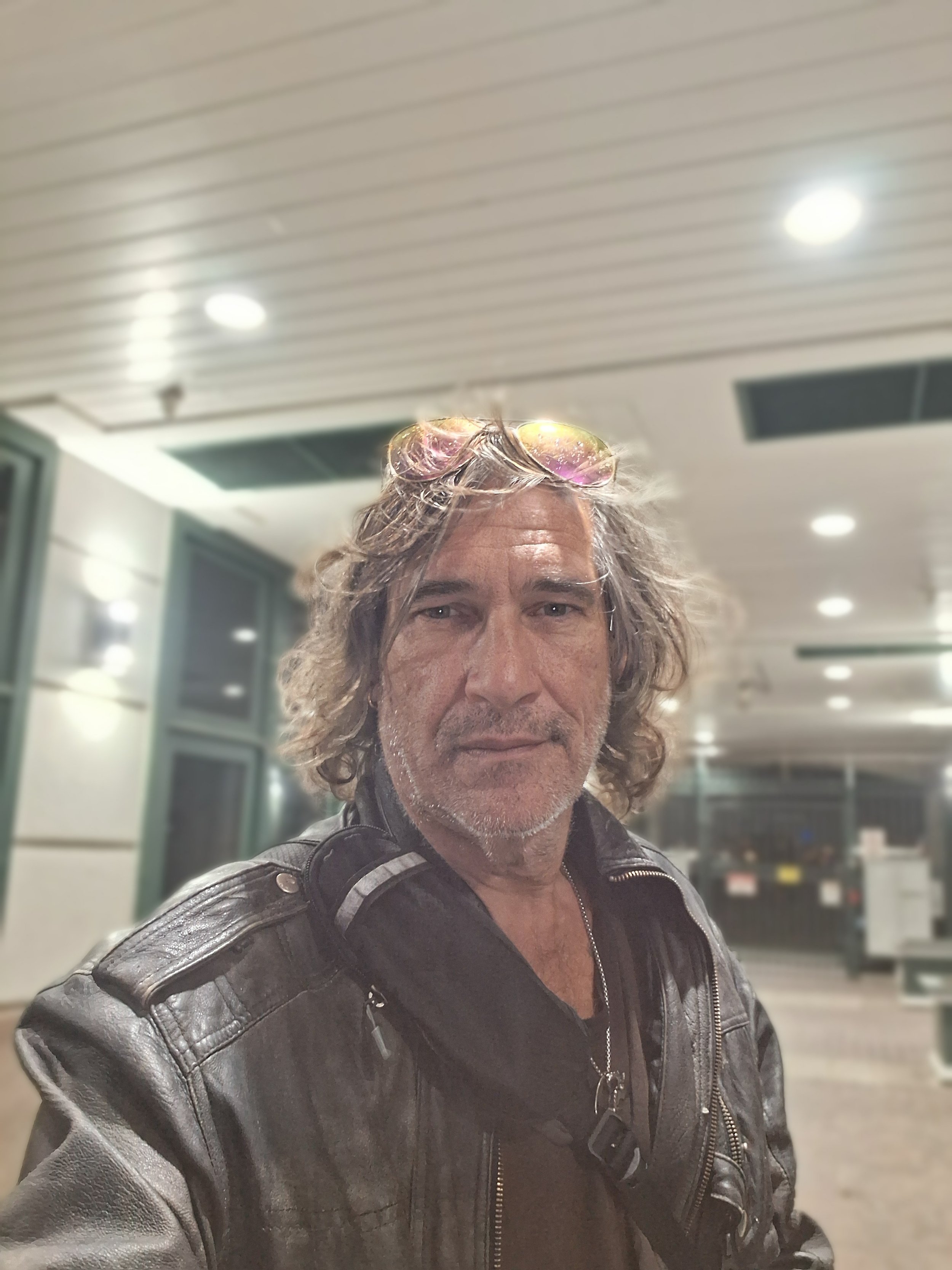 A man with curly hair and sunglasses on his head, wearing a leather jacket, taking a selfie indoors with a blurred background.