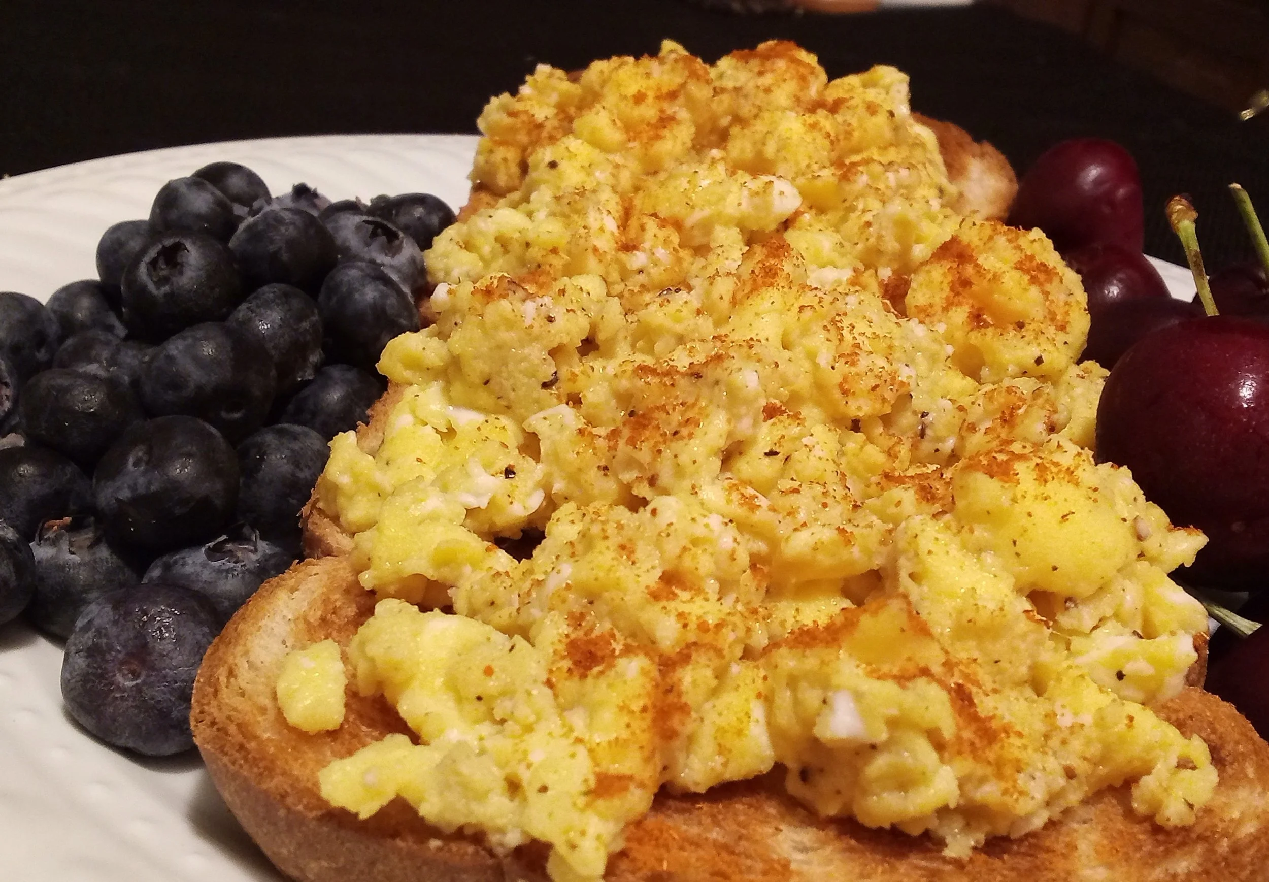 Close-up of scrambled eggs with toast, blueberries, and cherries on a white plate.