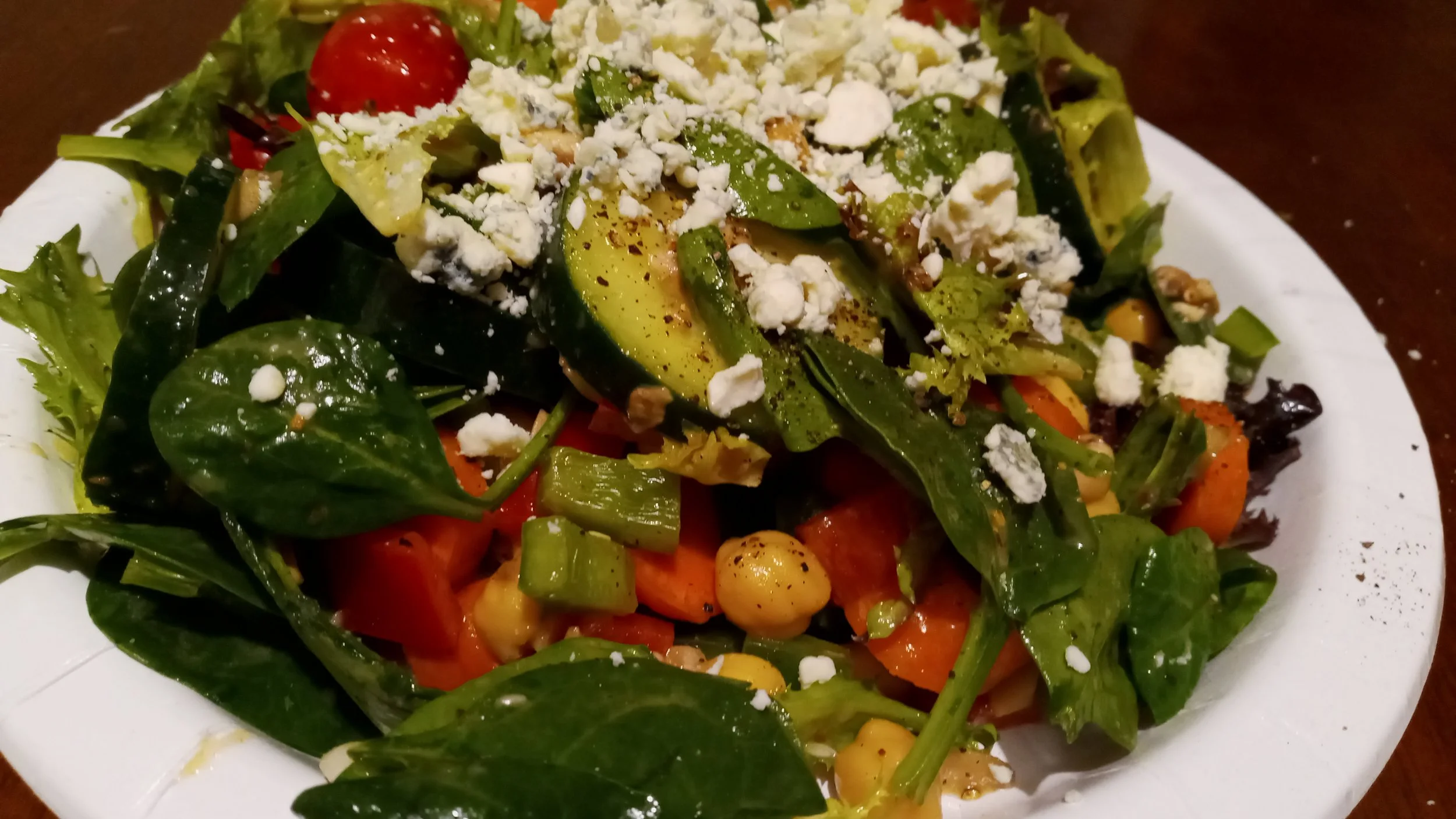 A plate of mixed green salad with avocado slices, cherry tomatoes, chopped bell peppers, chickpeas, and crumbled cheese.