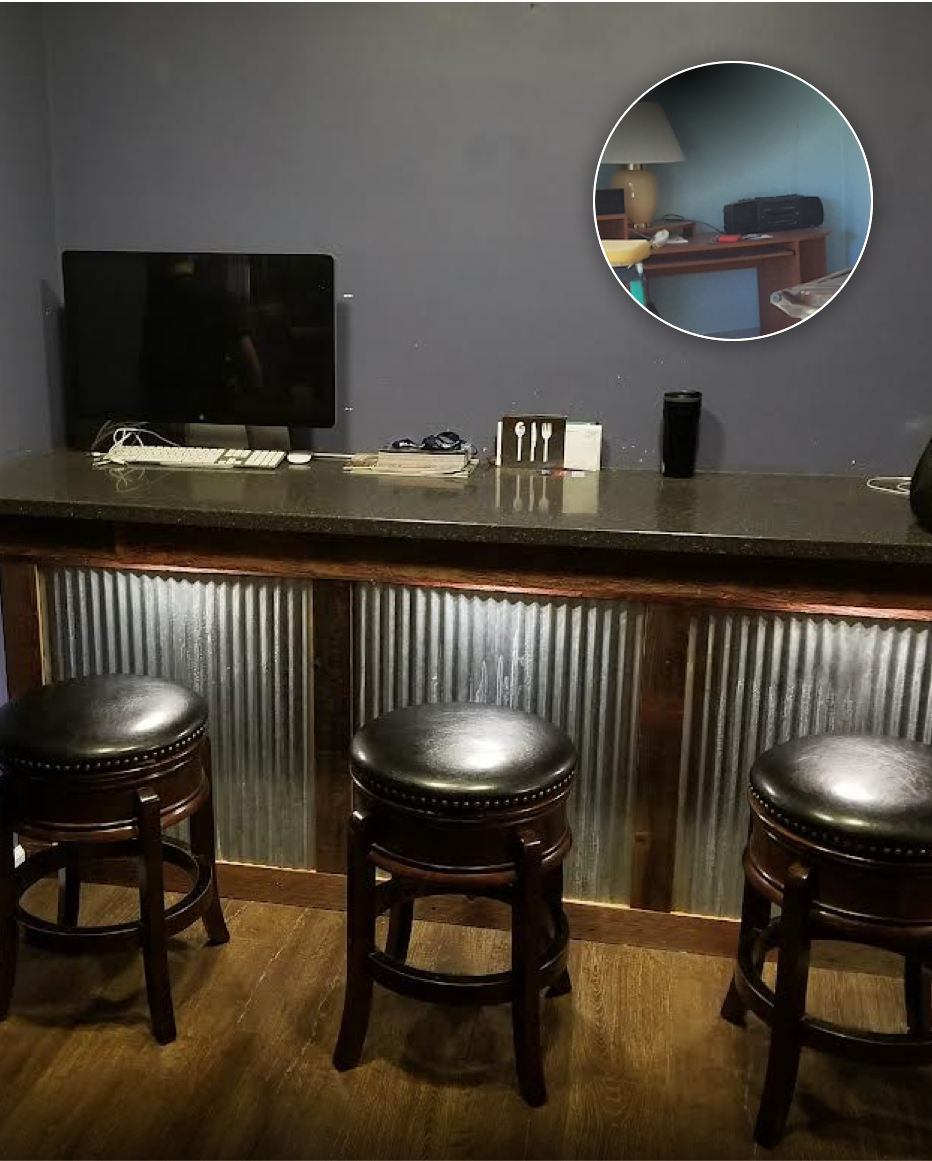 A bar area with three black leather stools, a black countertop, a computer monitor with keyboard and mouse, behind the counter are some small items, and a mirror reflecting a lamp and a printer.