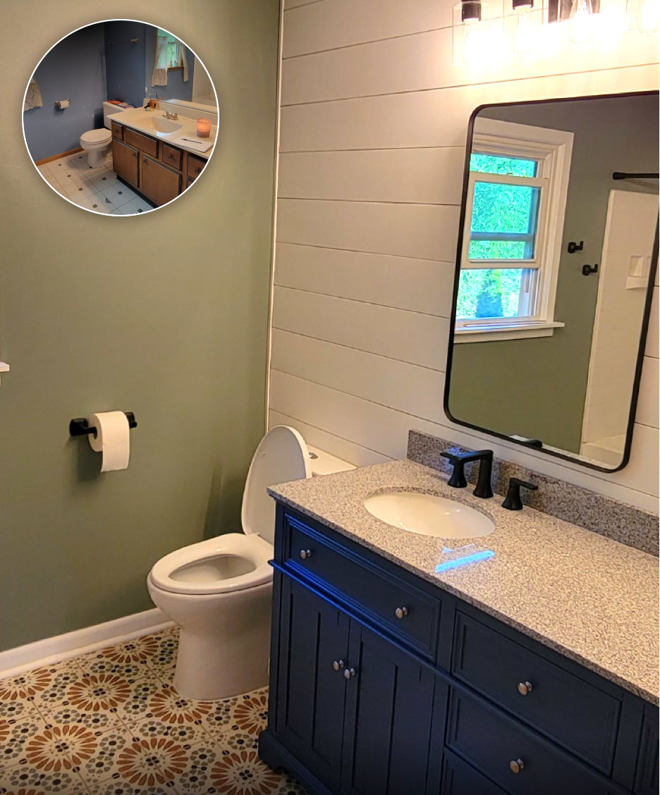 A bathroom with a gray wall, a white toilet, a blue cabinet with a granite countertop, a black faucet, a large rectangular mirror, a window with a view of green trees, and patterned floor tiles. The ceiling has a lighting fixture above the mirror.
