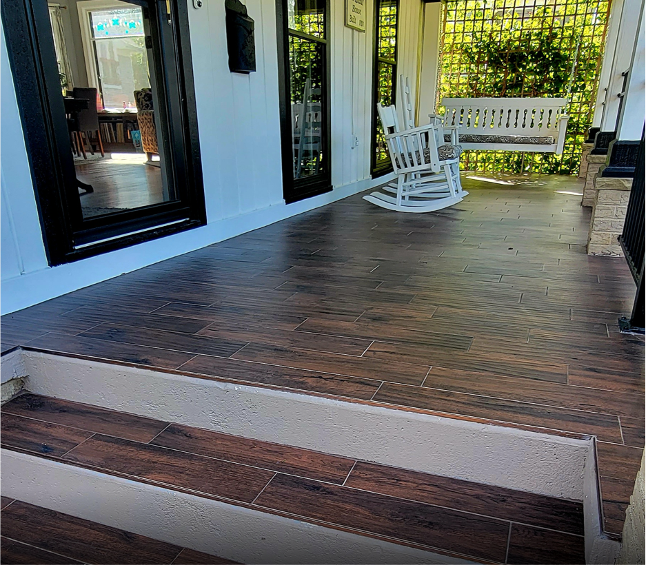 View of a screened porch with dark wood tile flooring, white wooden rocking chairs, a white porch swing, and greenery outside.