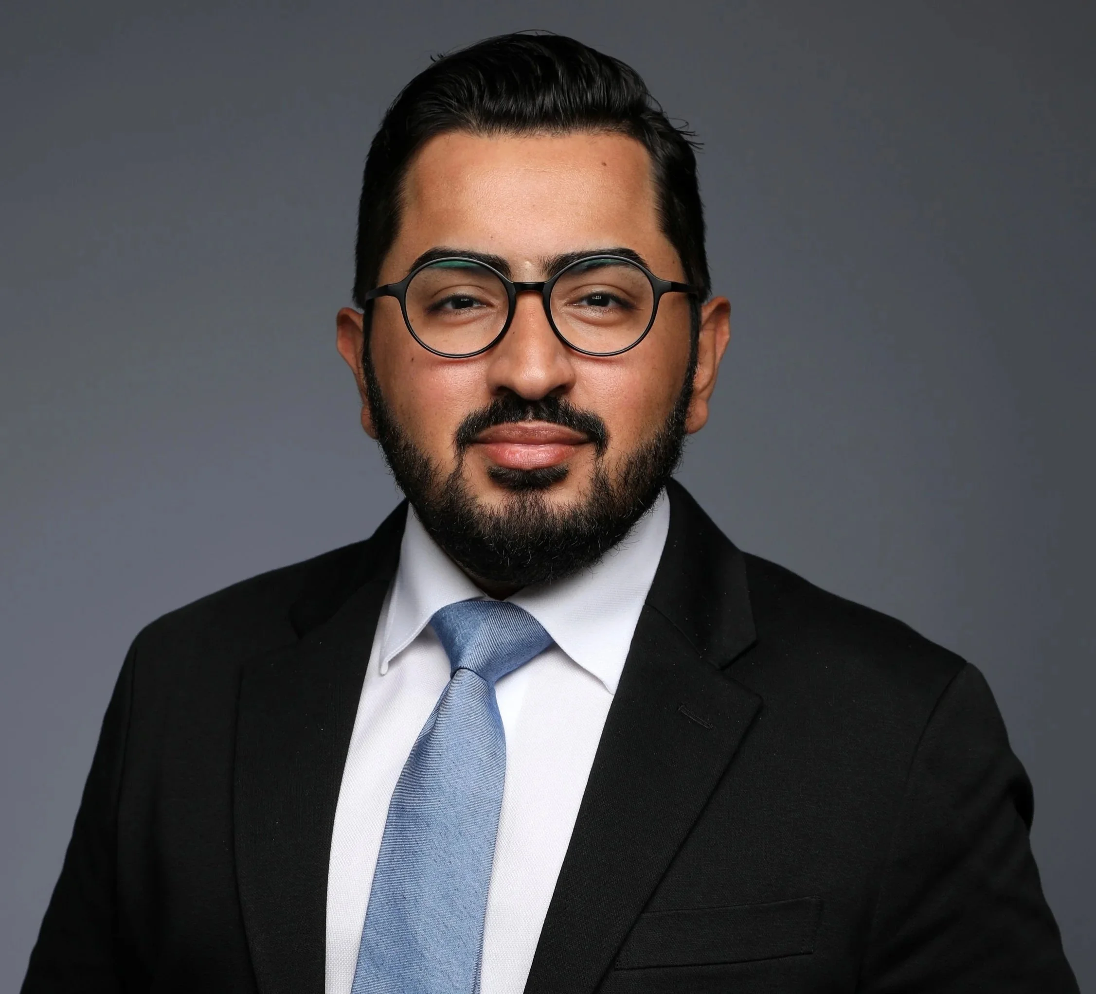 Professional headshot of a man wearing glasses, a black suit, white shirt, and blue tie against a gray background.