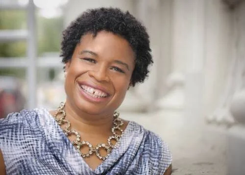 Portrait of a smiling woman with curly hair wearing a blue patterned blouse and a large pearl necklace.