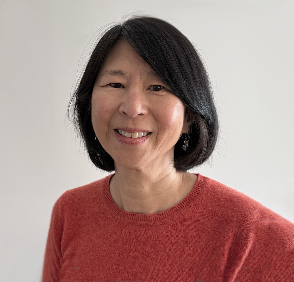 A woman with black hair smiling at the camera, wearing a red sweater and earrings, against a plain white background.