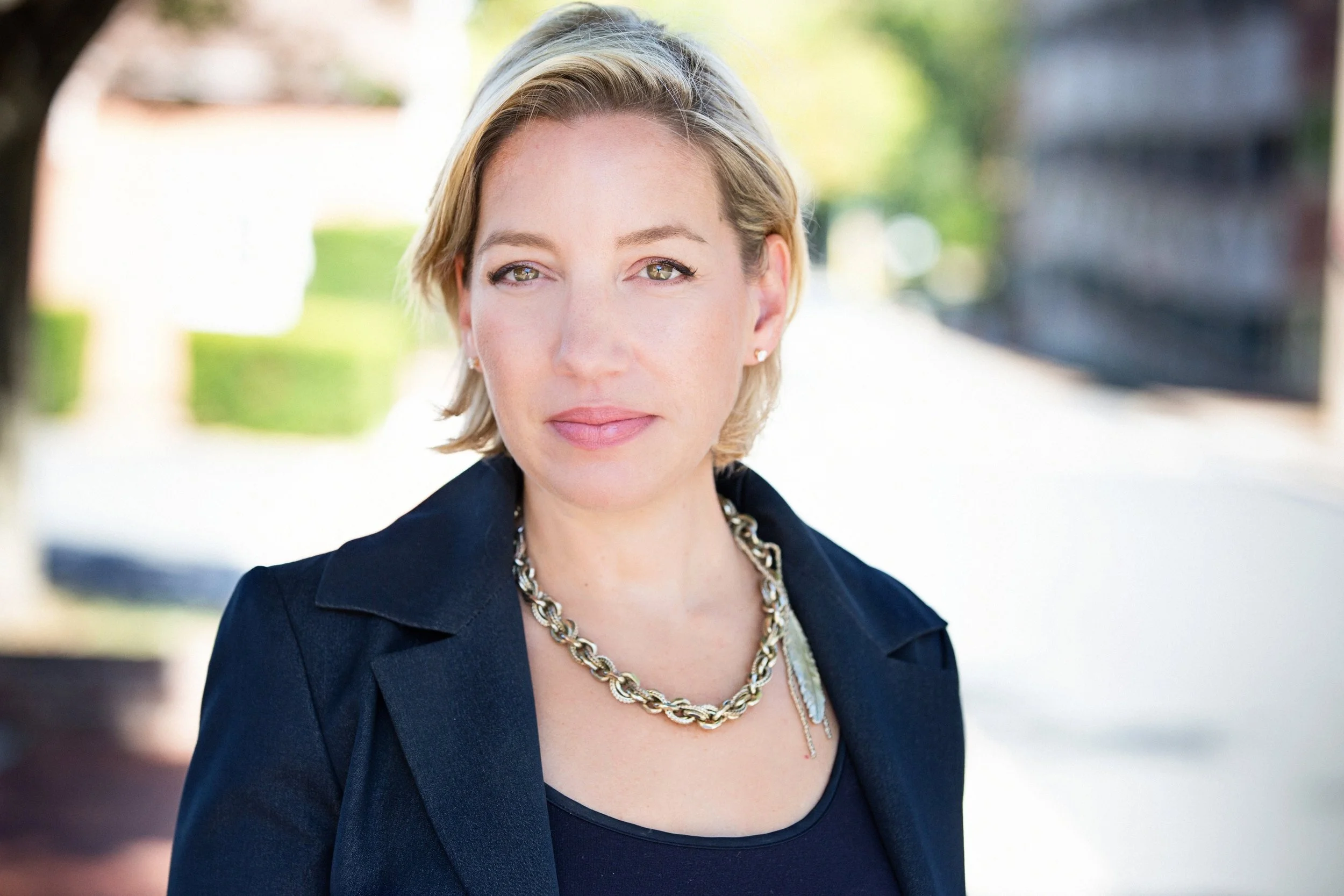 A woman with short blonde hair wearing a black blazer and a chunky gold chain necklace, standing outdoors with a blurred background of greenery and a building.