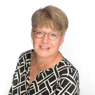 A middle-aged woman with short light brown hair, wearing glasses and a black and white patterned blouse, smiling at the camera against a white background.