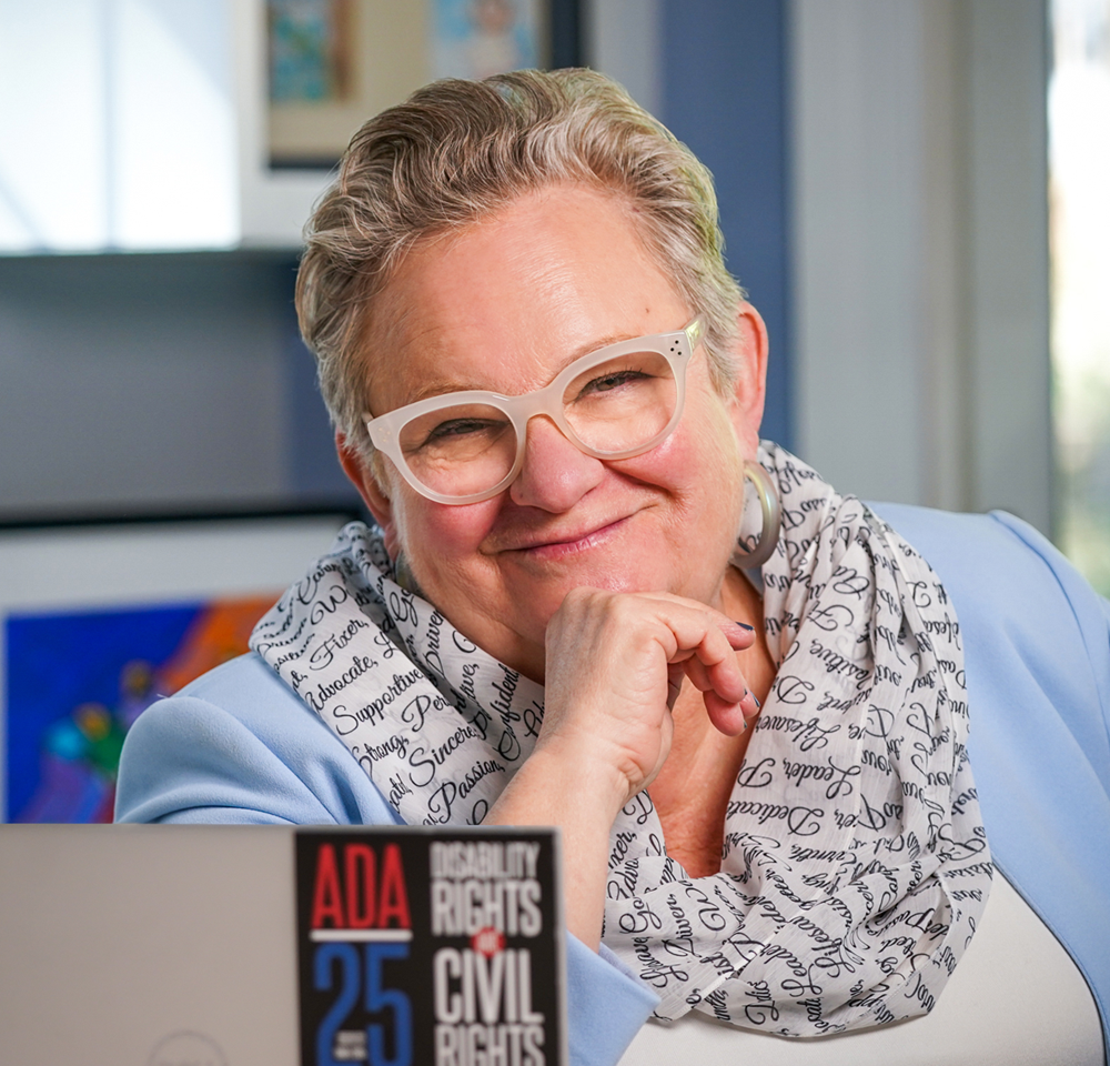 A smiling woman with short gray hair wearing glasses and a light blue blazer, sitting at a desk with a sign about disability rights in the foreground.
