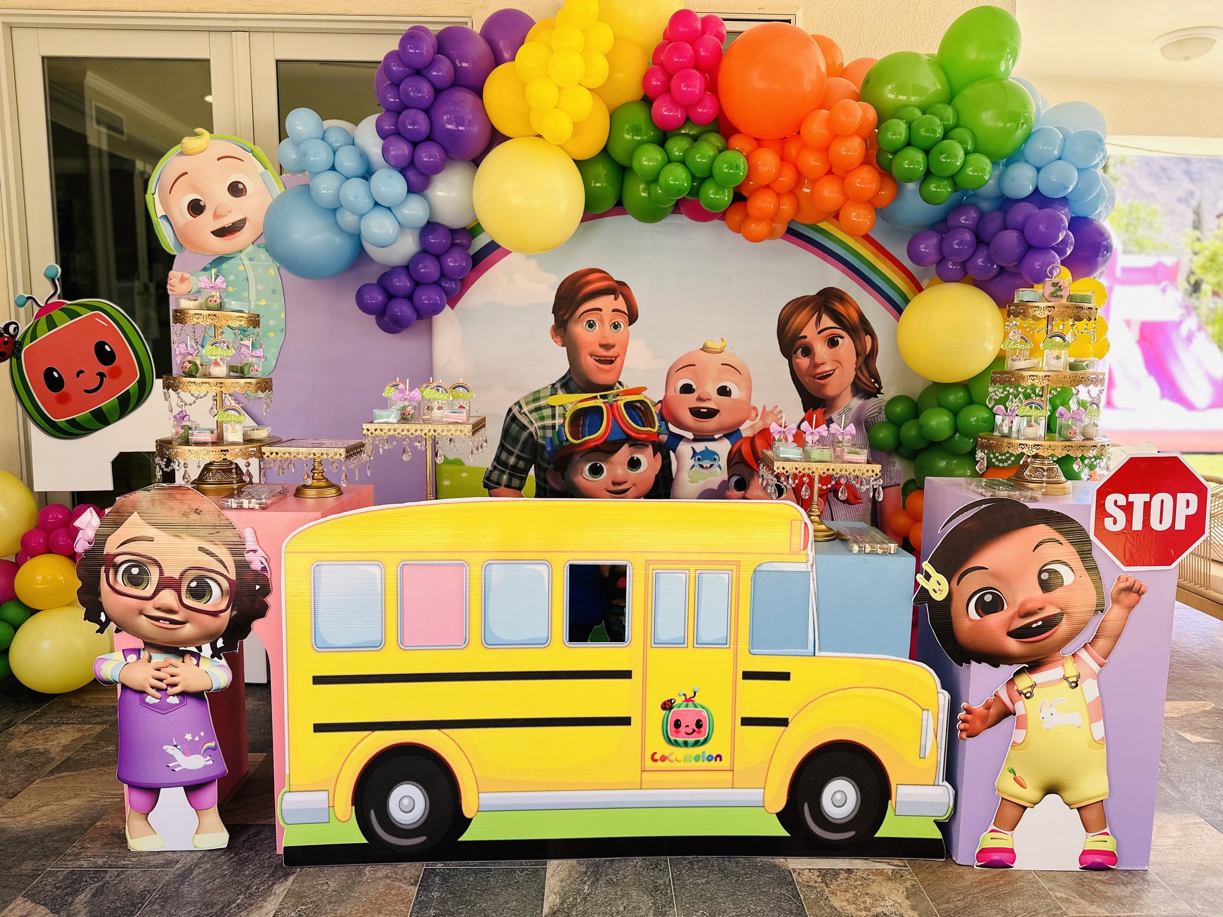 Colorful children's birthday party with a backdrop of cartoon characters, balloons, and a cutout of a yellow school bus from the show Coco Melon, featuring characters from the animated series.