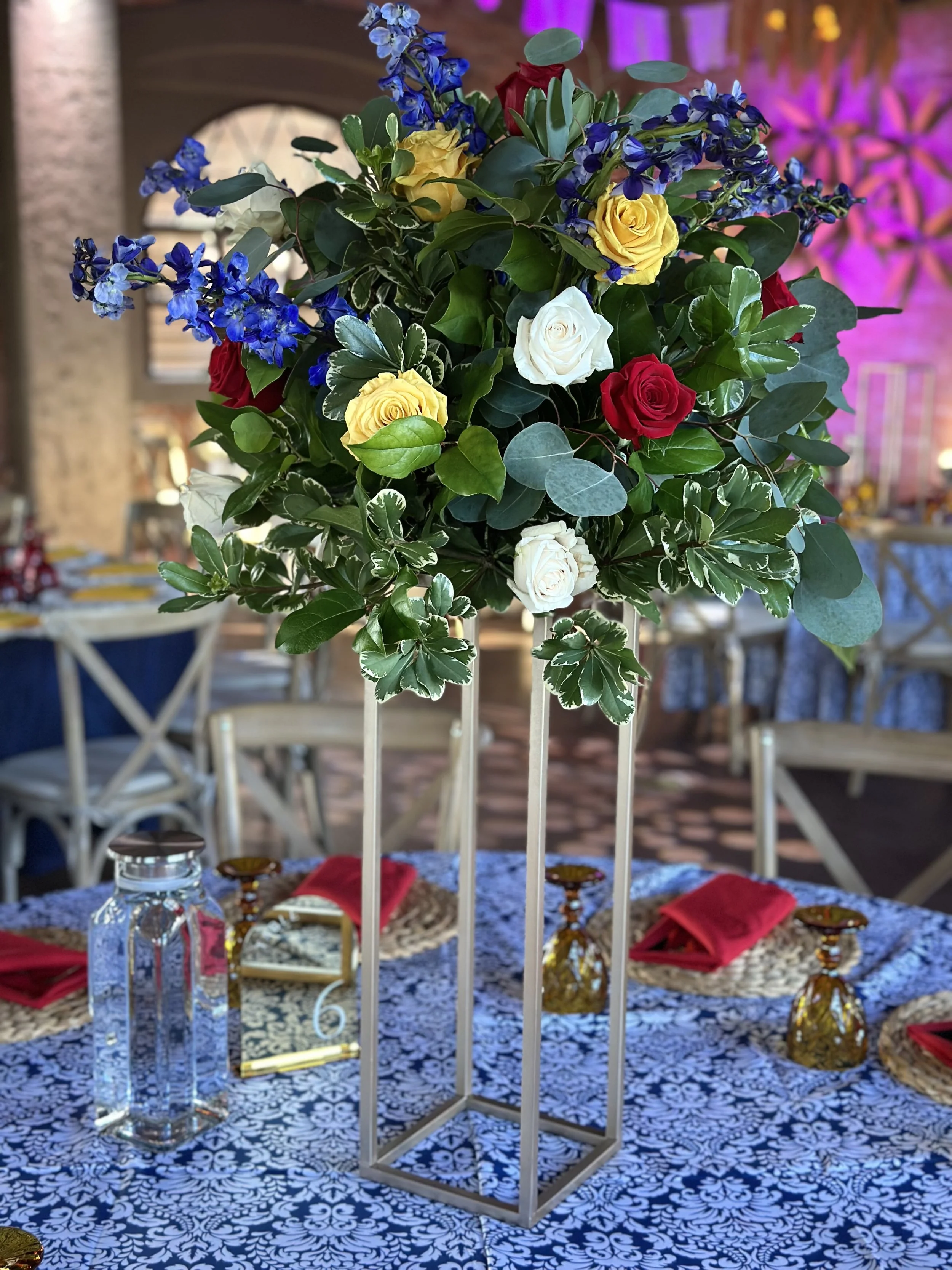 A tall floral centerpiece with yellow, white, and red roses, and blue delphiniums.