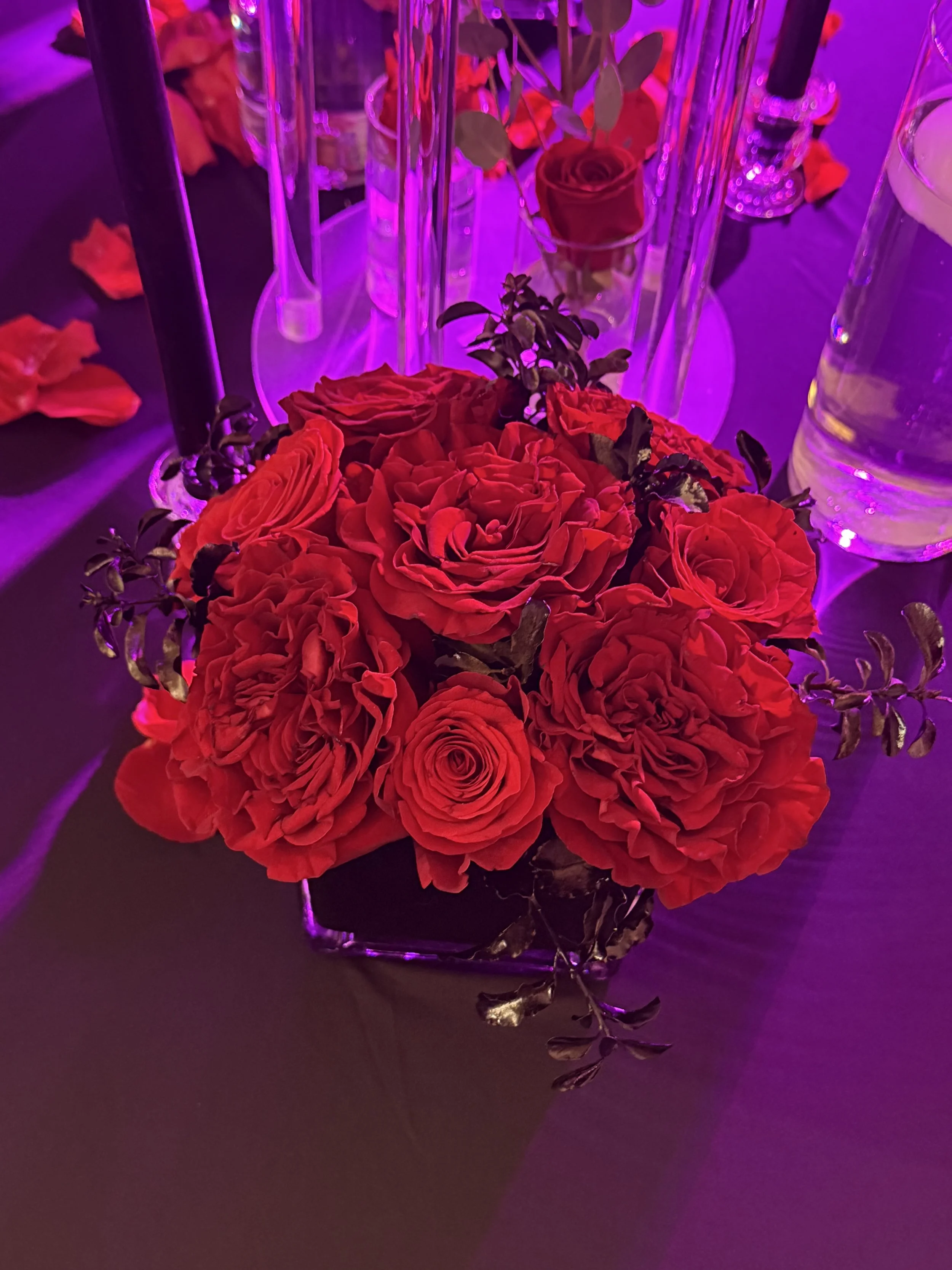 A floral centerpiece with red roses and other red flowers in a square glass vase, surrounded by a table setting with candles, a water bottle, and rose petals, under purple lighting.