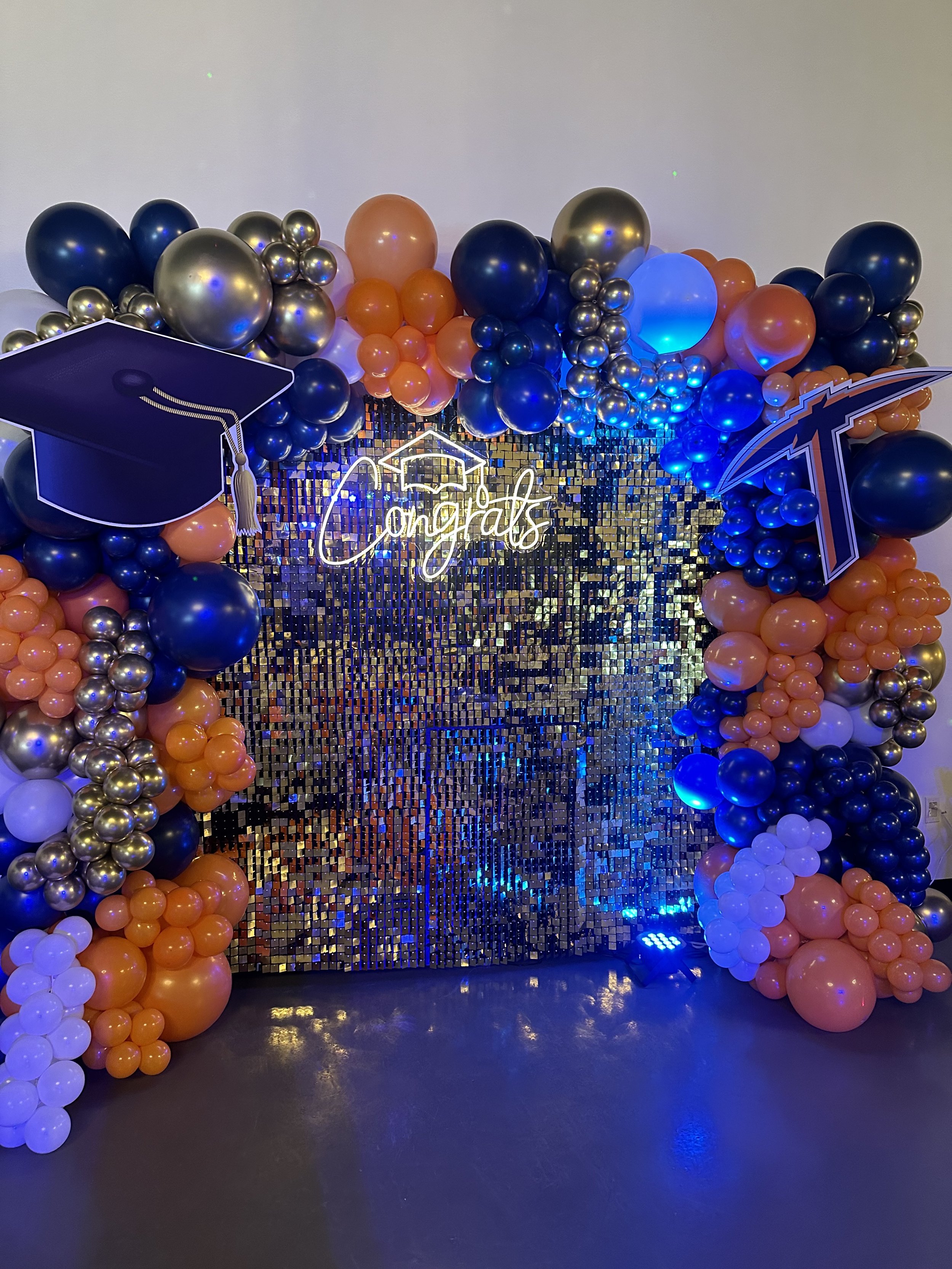 Graduation-themed balloon arch with a neon 'congrats' sign, a diploma cap, and a large graduation cap cutout, golden and blue balloons, a blue light, and a shiny mosaic backdrop.