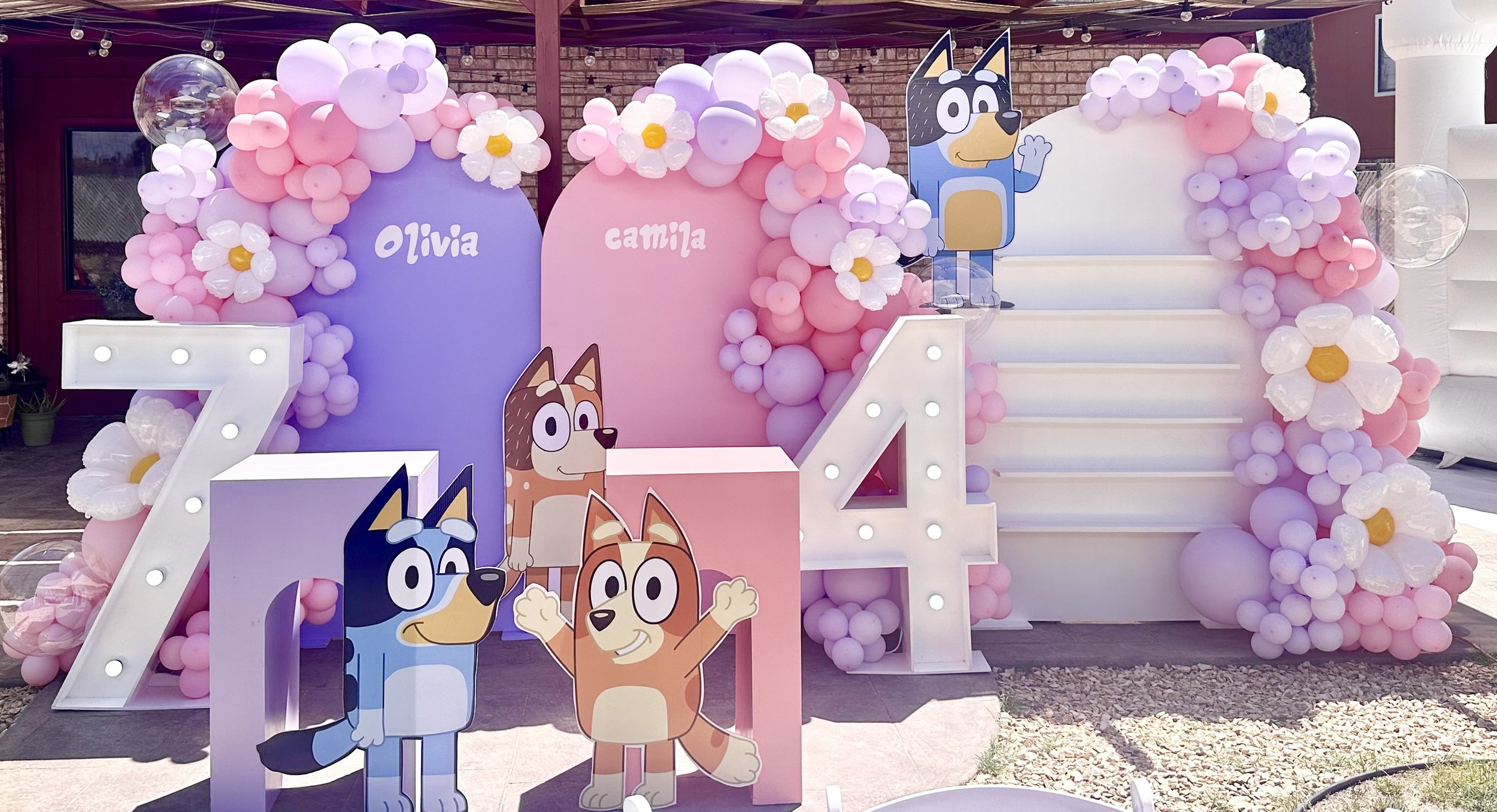 Decorative birthday setup with large helium balloons, large illuminated numbers '7' and '4', cartoon animal characters, and floral balloon arrangements, celebrating Olivia and Camila's birthday.