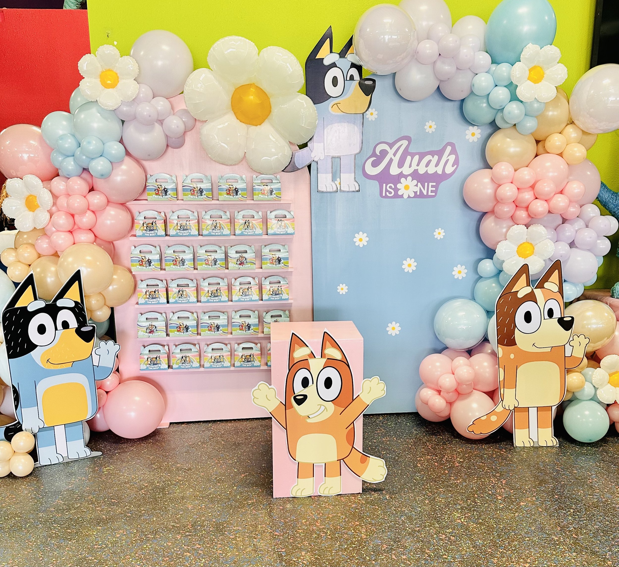 Decorative birthday party setup with cartoon dog characters and pastel-colored balloons. The backdrop displays the message 'Auah is One' with images of cartoon dogs. There are various cartoon dog cutouts and boxes with cartoon dog illustrations, like