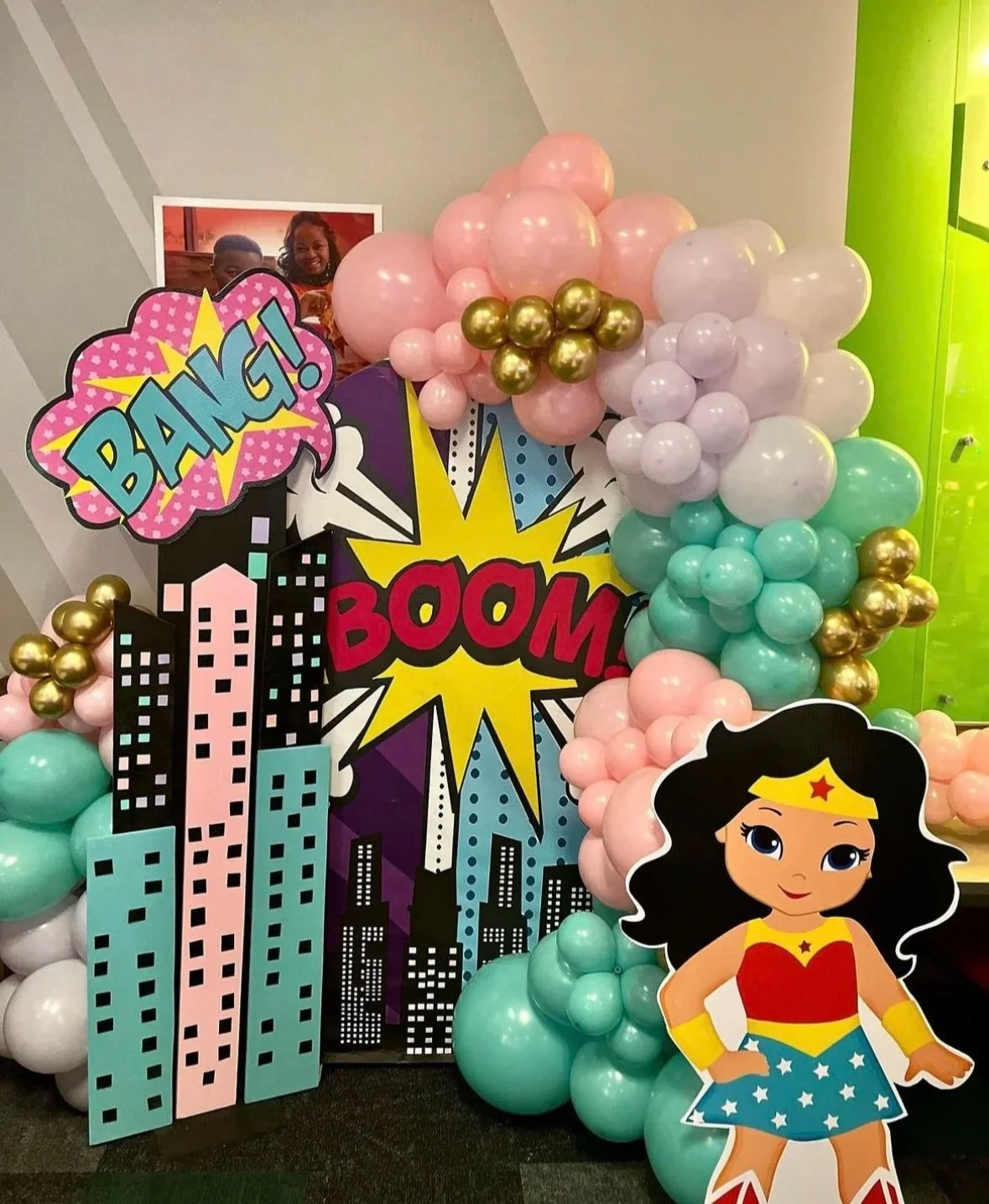 Decorative superhero-themed party backdrop featuring balloons, comic book-style sound effects like 'BANG!' and 'BOOM!', a cartoon Wonder Woman cutout, and a photo of smiling children in the background.