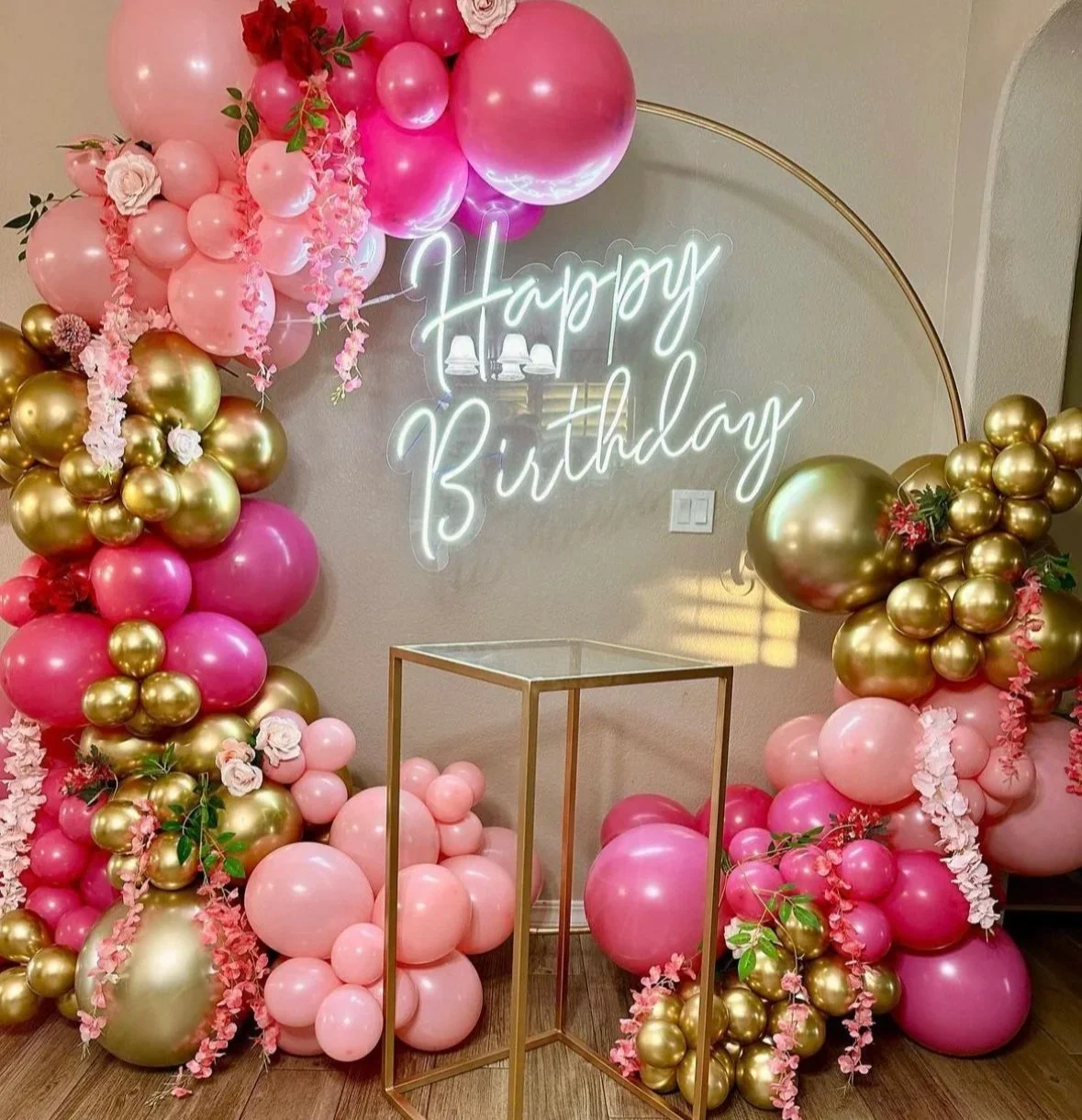 Balloon arrangement with pink, gold, and light pink balloons, pink flowers, and a glowing neon sign that reads 'Happy Birthday'.