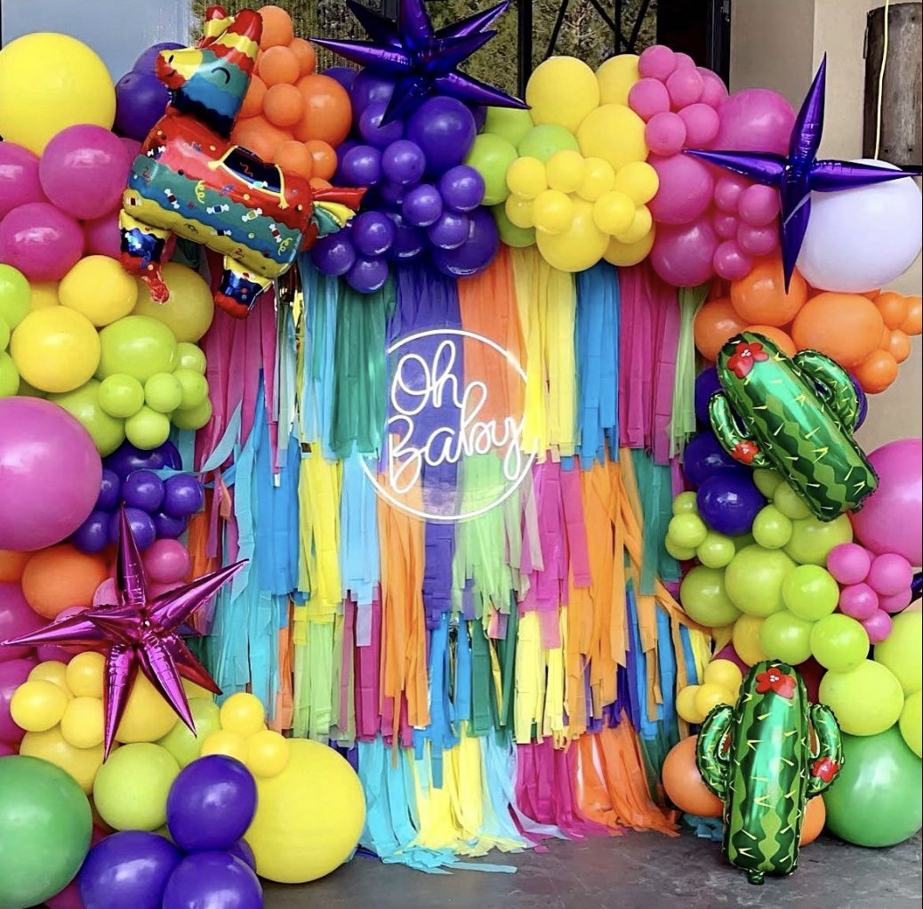 Colorful balloon wall with rainbow-colored paper streamers hanging in front, featuring a sign that reads 'Oh Baby' in the center. The balloons include pink, yellow, green, purple, and orange, some shaped like cacti, with star-shaped metallic balloons