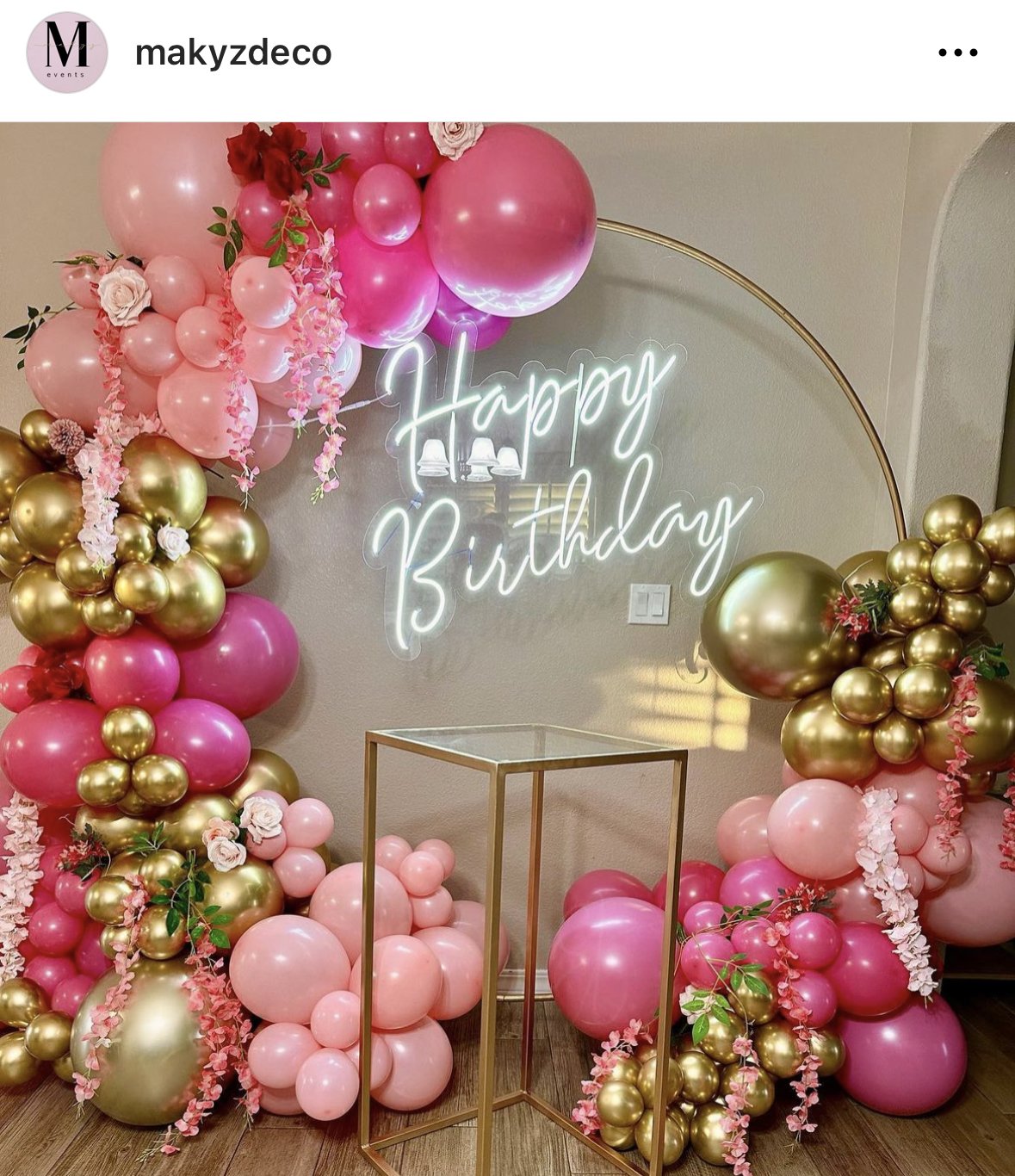 Pink and gold balloon arch with flowers, neon 'Happy Birthday' sign, and a glass display table.