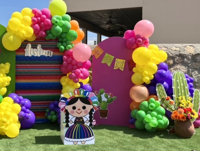 Colorful birthday party backdrop with balloons, a pink backdrop with yellow, green, purple, and pink balloons, a paper cutout of a girl in traditional Mexican dress, cactus cutouts, and vibrant decorations.