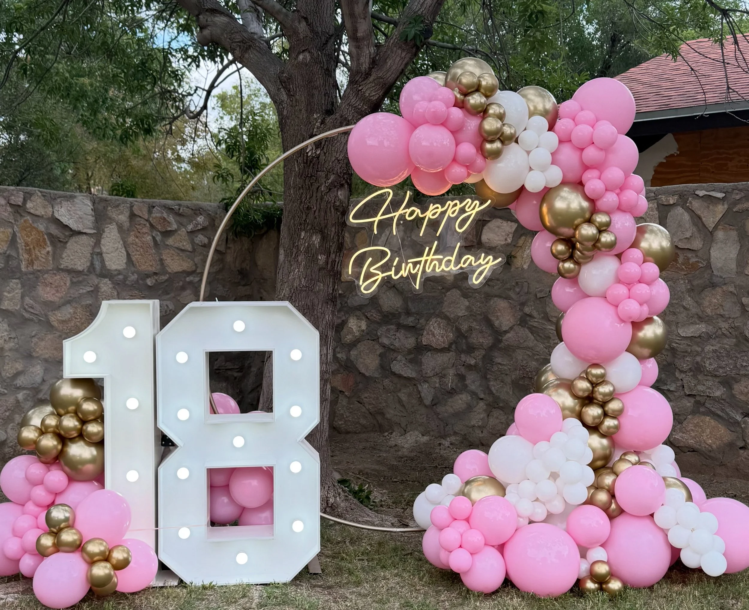 Birthday outdoor celebration setup with pink, white, and gold balloons, a large illuminated number 18, and a neon sign that says "Happy Birthday" near a tree and stone wall.