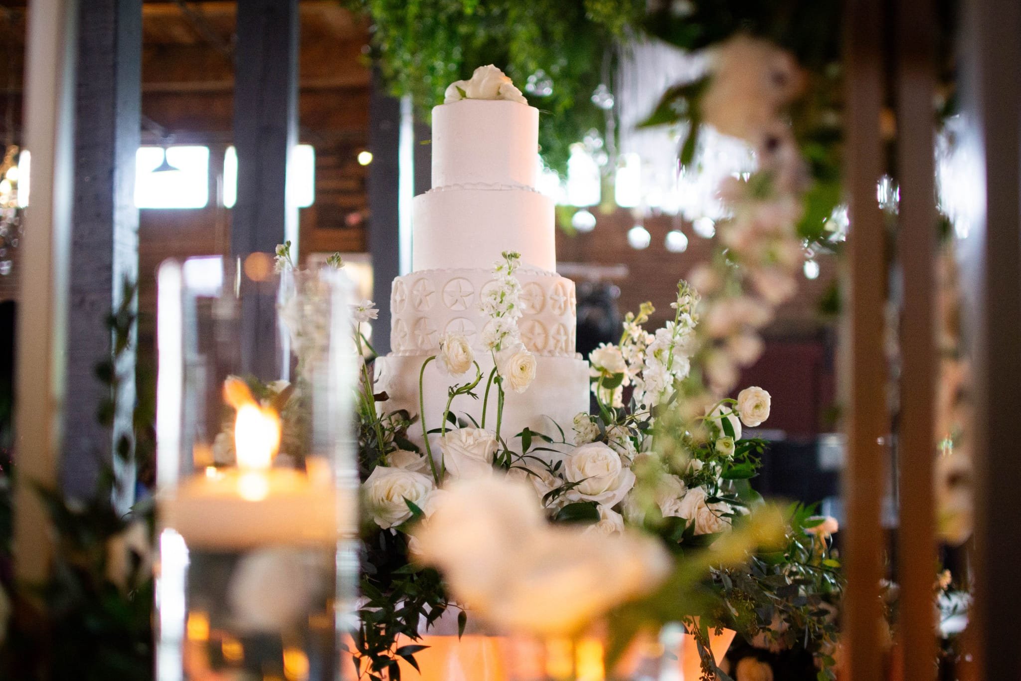 A tall, white, multi-tiered wedding cake decorated with white flowers and surrounded by white roses and greenery, on a table with candles in the foreground, in a venue with large windows and wooden accents.