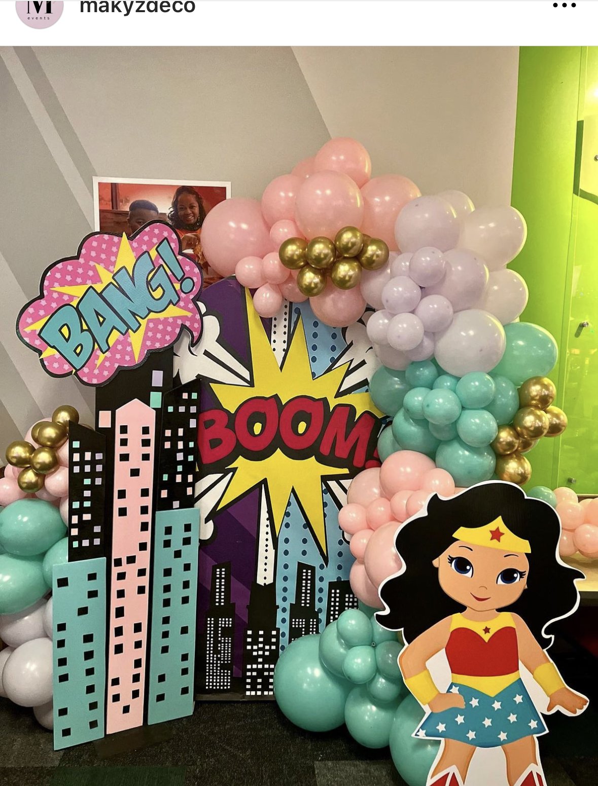 Celebration decoration featuring a superhero comic book theme with balloons, a large cartoon Wonder Woman figure, and colorful comic-style signs with words like 'BANG!' and 'BOOM!'.