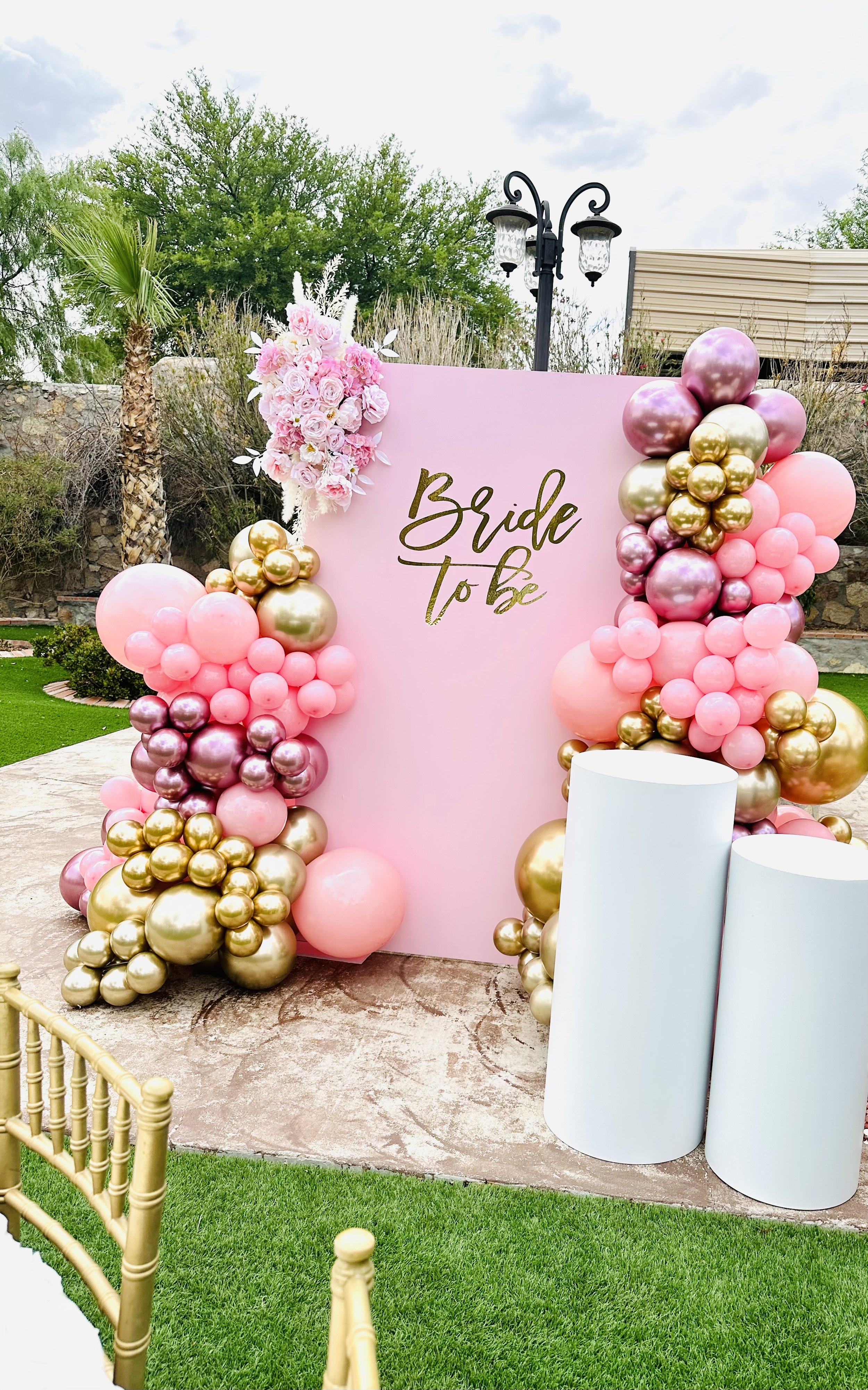 Decorative pink backdrop with the words 'Bride to be' in gold, surrounded by pink, gold, and purple balloons and a floral arrangement, set outdoors with palm trees and a cloudy sky in the background.