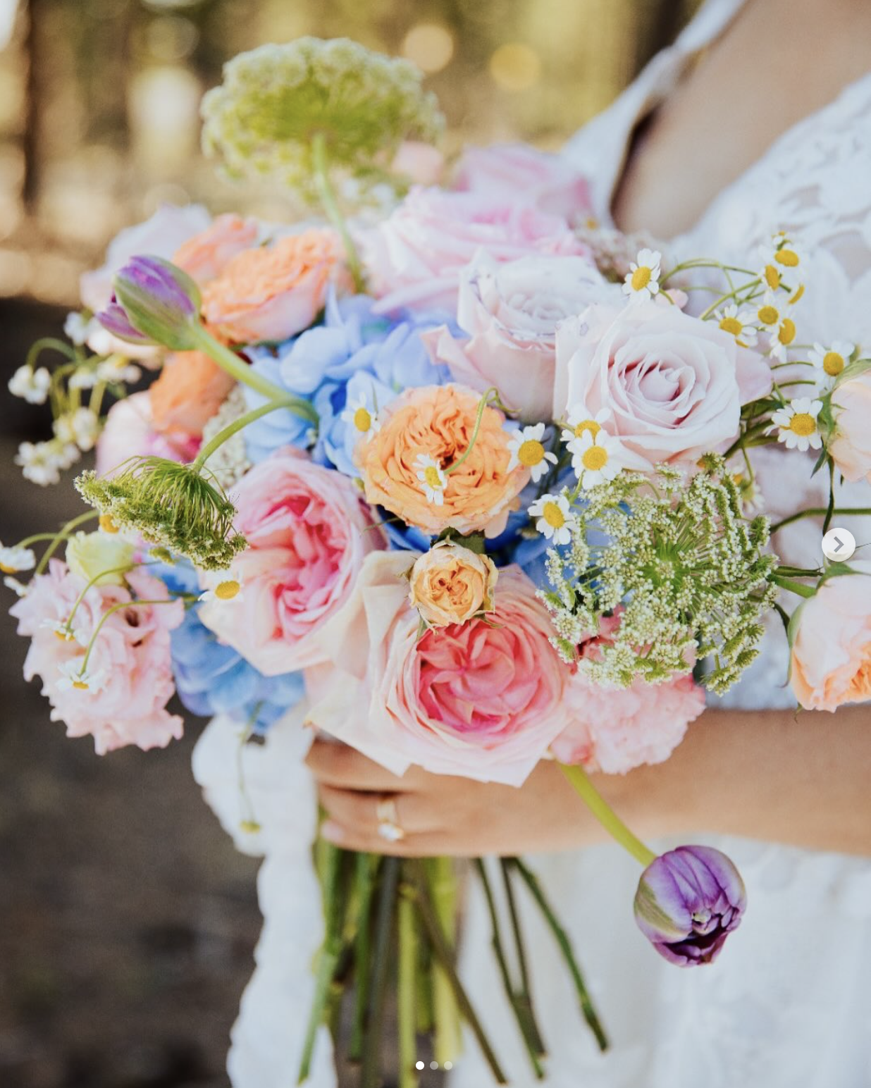 A person holding a colorful bouquet of flowers including pink roses, peach ranunculus, blue hydrangeas, and small white daisies.