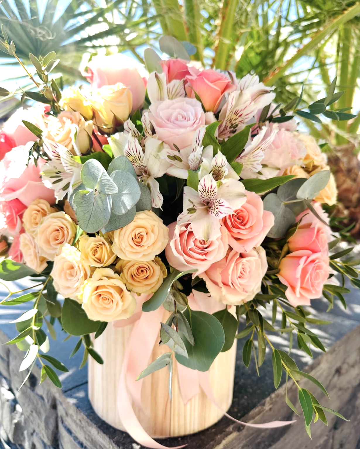 Ramo de flores con rosas rosadas, alstroemerias blancas con rojo, y hojas verdes en jarrón de madera.