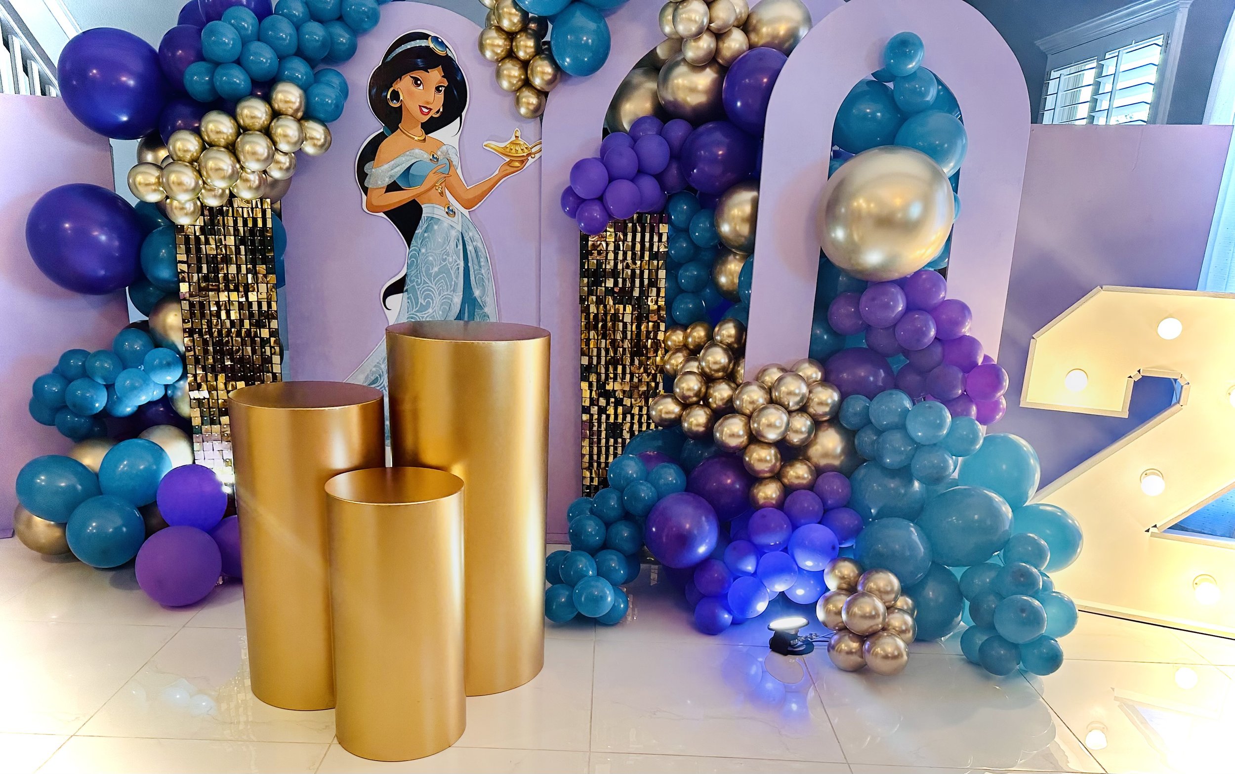 Party decoration featuring a cartoon princess with a purple, blue, and gold balloon arch, gold cylinders, and a large illuminated number two, with a backdrop of purple and blue balloons and a princess-themed poster.