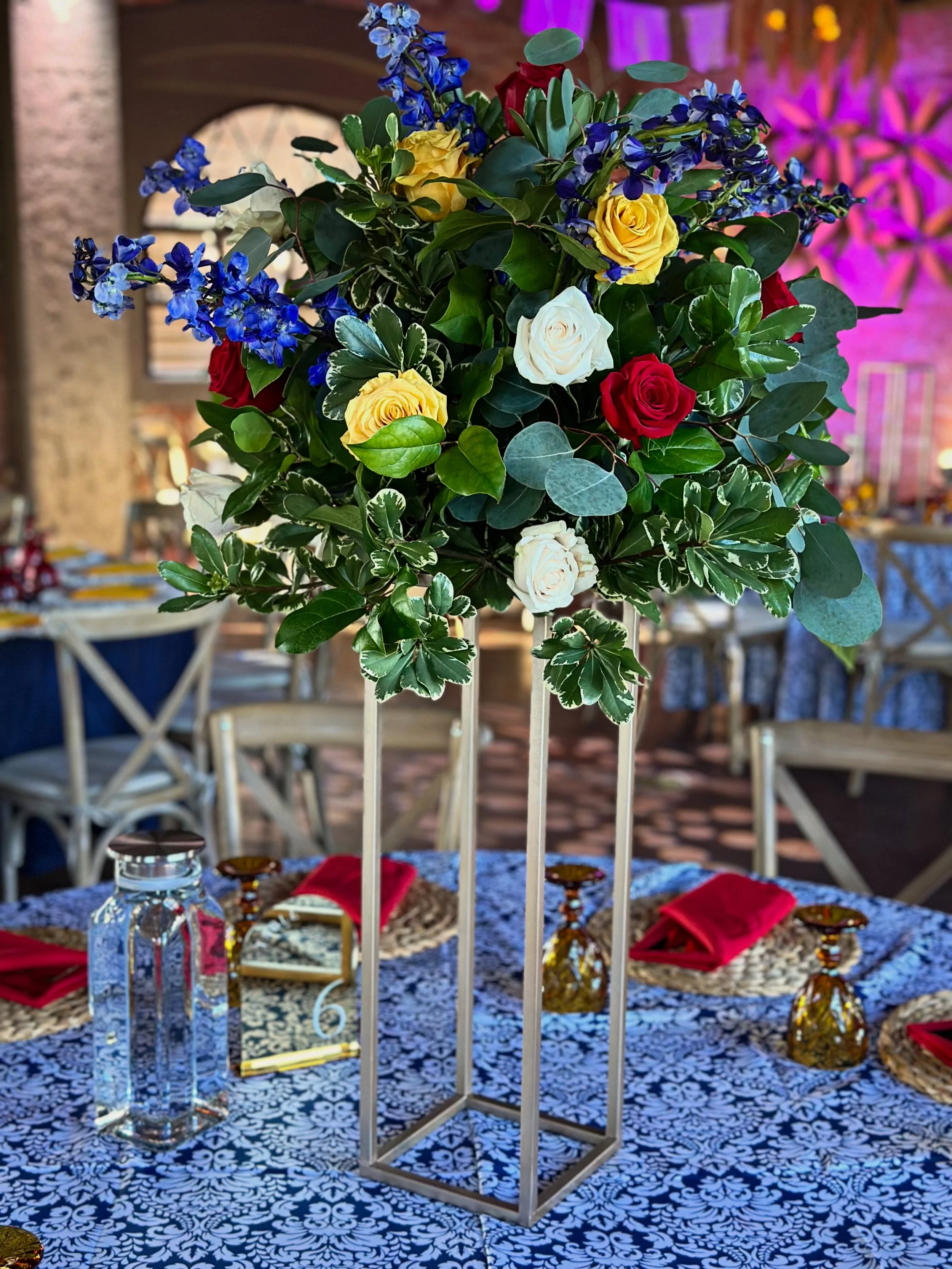 A tall floral centerpiece with yellow, white, red roses, and blue delphiniums.