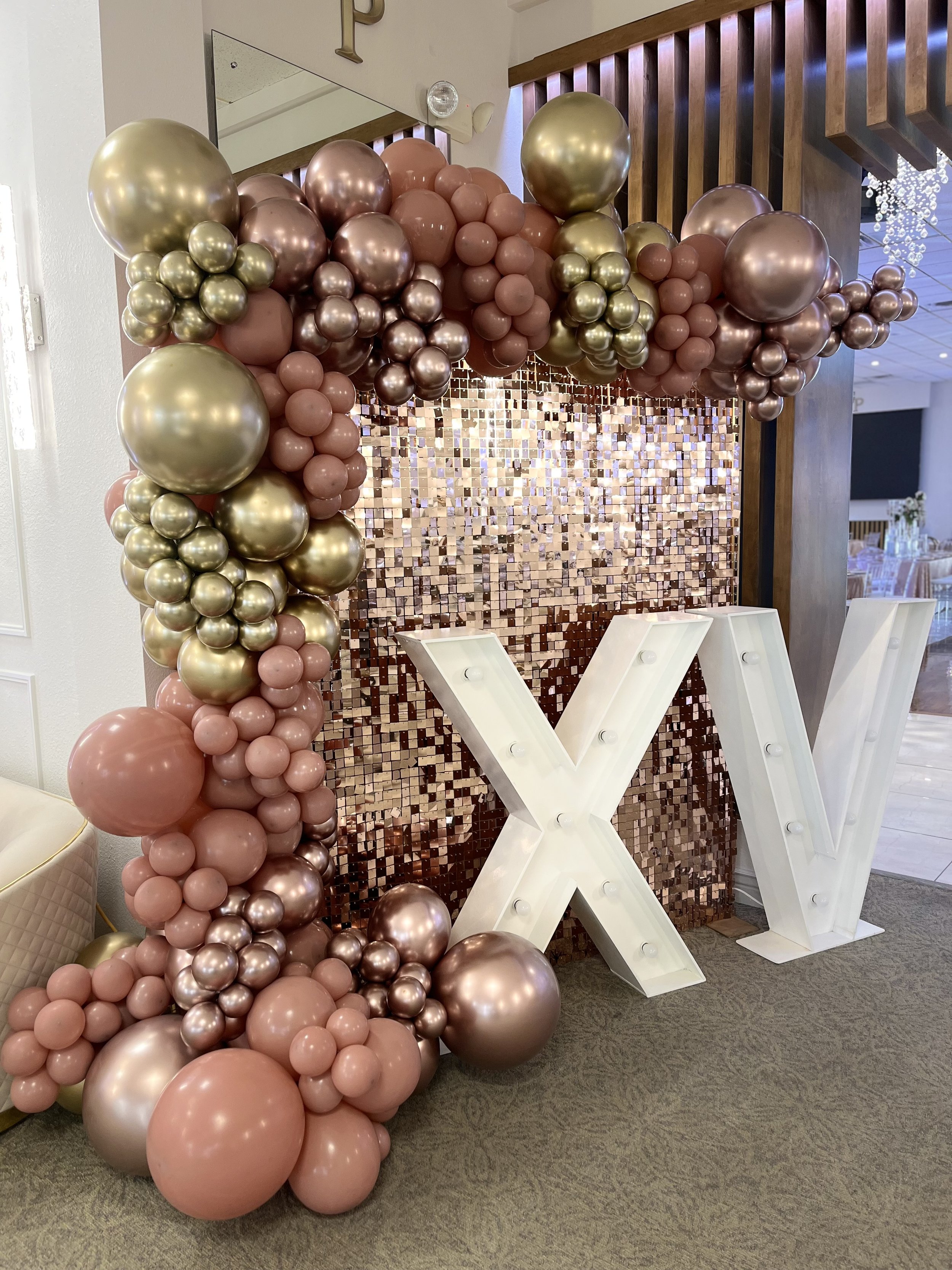 Decorative balloon arch in shades of rose, pink, and gold with large illuminated letters forming the letter 'X' below a shimmering pink sequin backdrop.