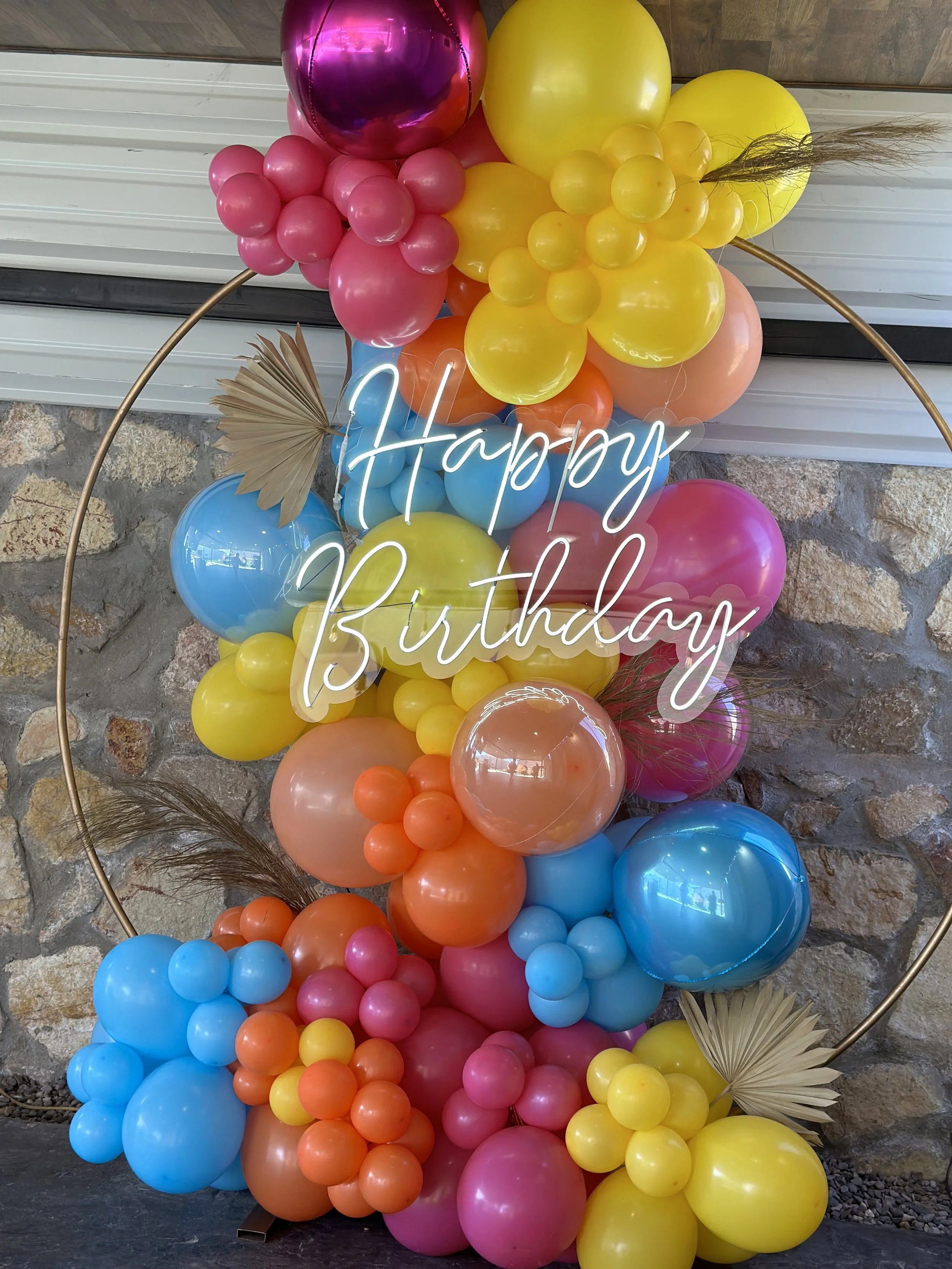 Colorful birthday balloon arrangement with pink, yellow, orange, blue, and purple balloons, gold pampas grass, and a neon sign reading 'Happy Birthday' in white cursive text, set against a stone wall background.
