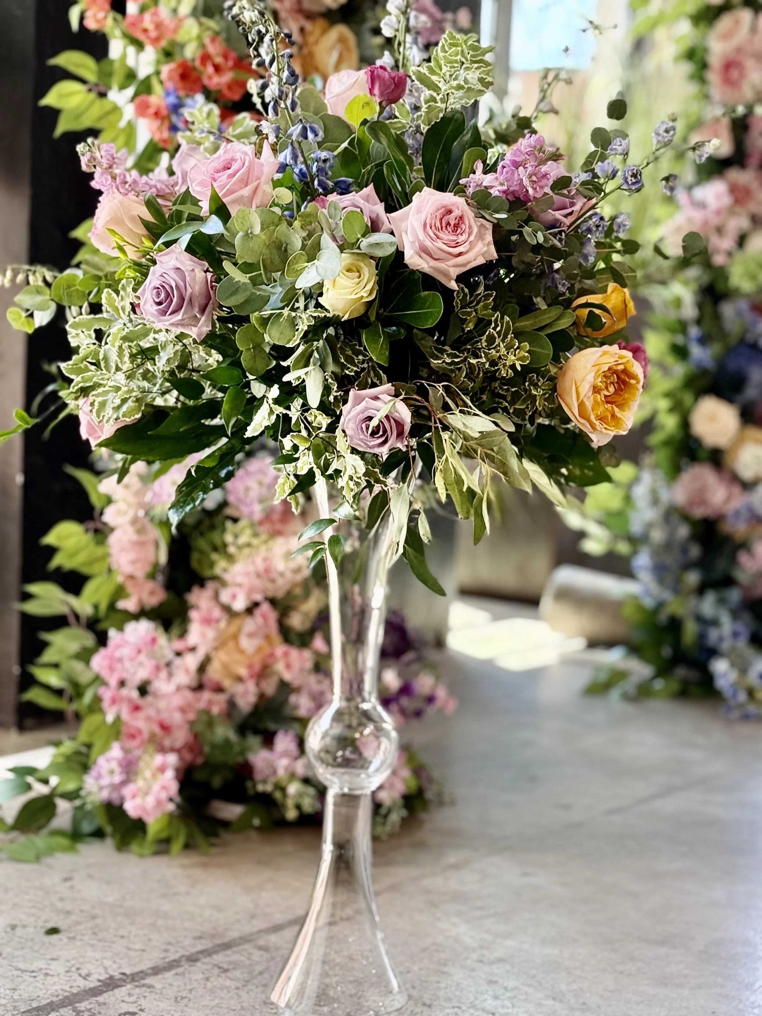 A glass vase holding a colorful bouquet of pink, purple, and yellow roses, with green foliage, on a table with more floral arrangements in the background.