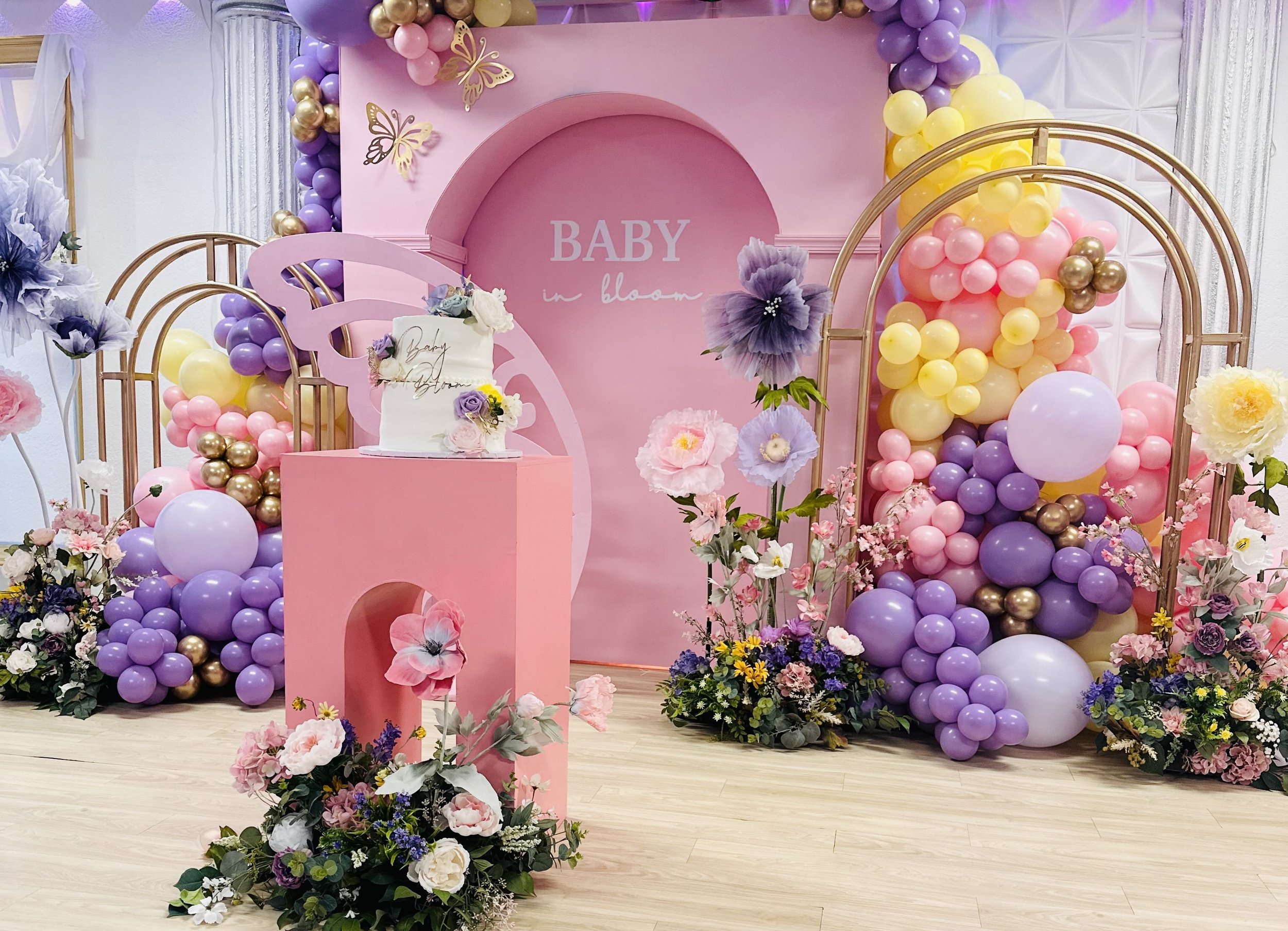 Decorative baby shower setup with pink and purple balloons, flowers, and a cake on a pink stand, featuring the words 'Baby in Bloom' on the backdrop.