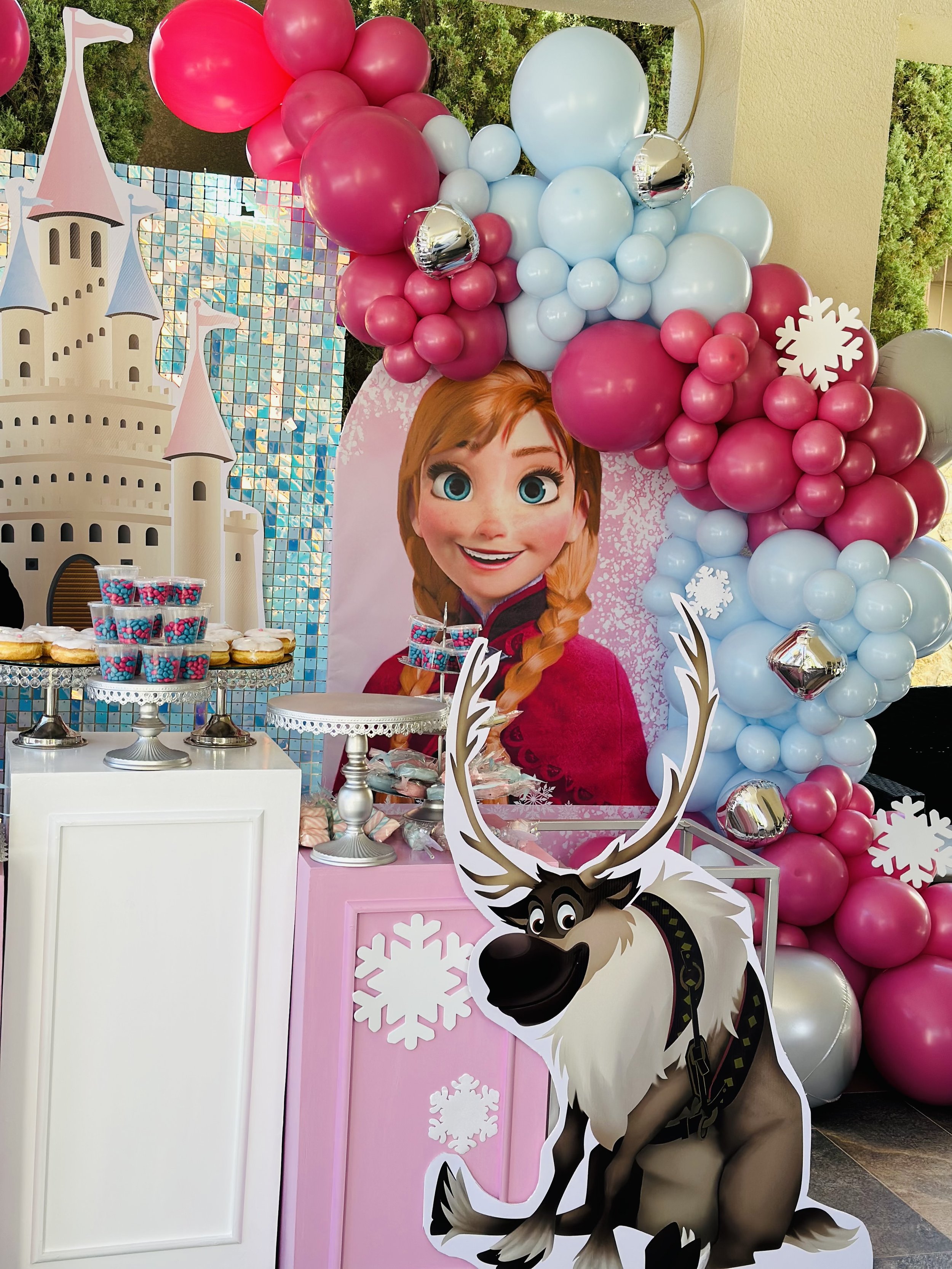 Disney Frozen themed birthday party display featuring a large cardboard cutout of Anna, snowflake decorations, a balloon arrangement in shades of pink, white, and silver, a cartoon reindeer figure, and a table with cupcakes and treats.