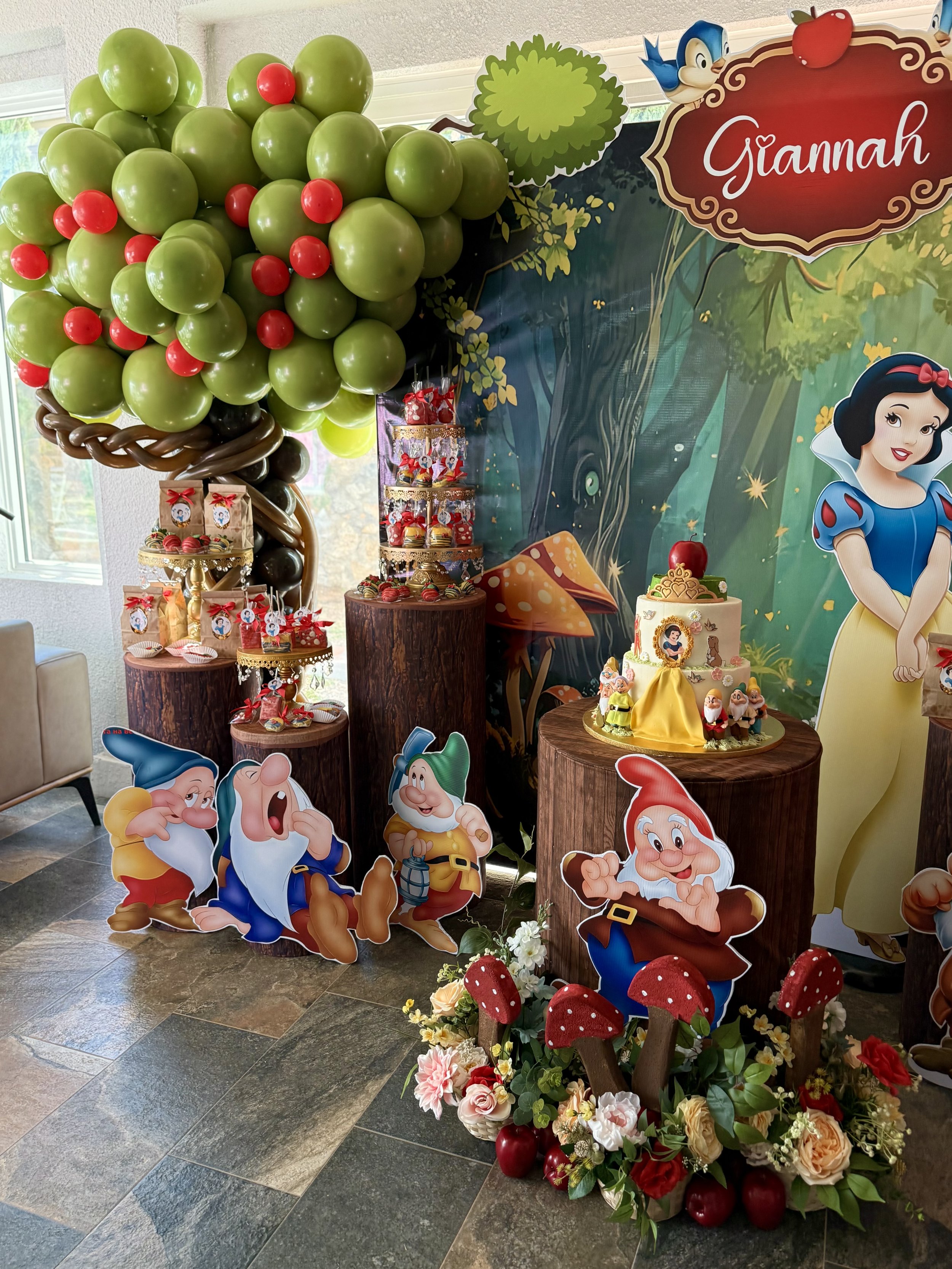 Decorations for a Snow White themed birthday party featuring a balloon apple tree, illustrated characters from Snow White, a tiered cake with Snow White and dwarfs figurines, and various themed treats and floral arrangements.