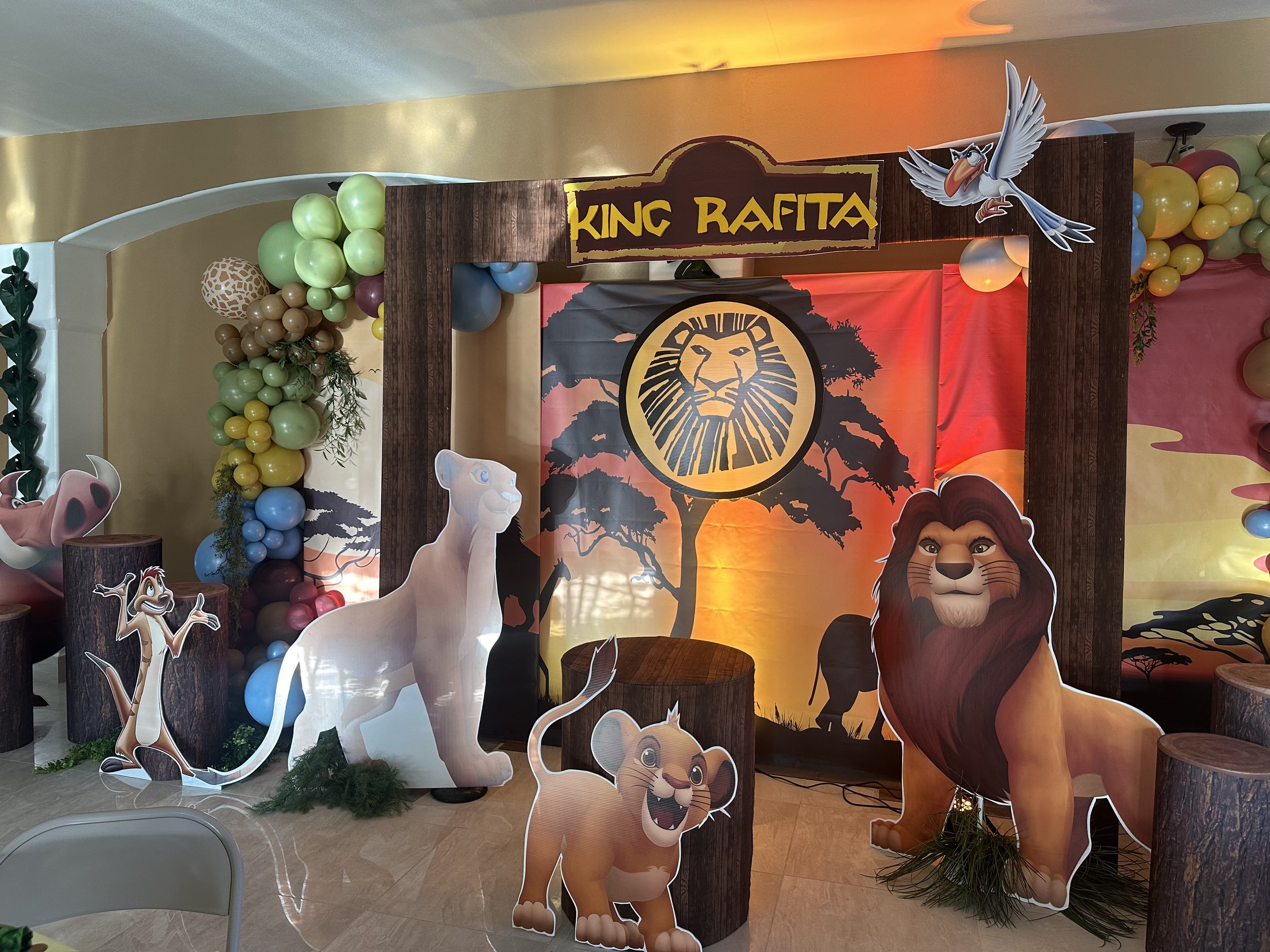 Decorative jungle-themed setup with cutouts of lions, a cheetah, a meerkat, and balloons, featuring a backdrop with a tree silhouette and the words "King Rafita" on a sign.
