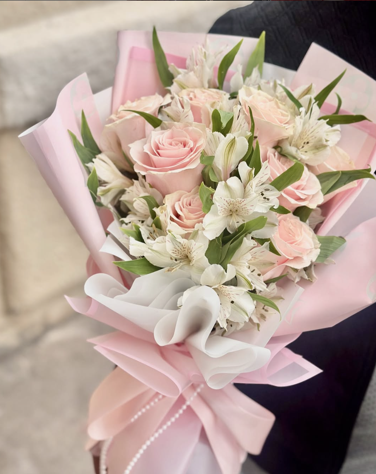 Ramo de flores con rosas rosas y lirios blancos, envuelto en papel rosa y blanco con lazo rosado.