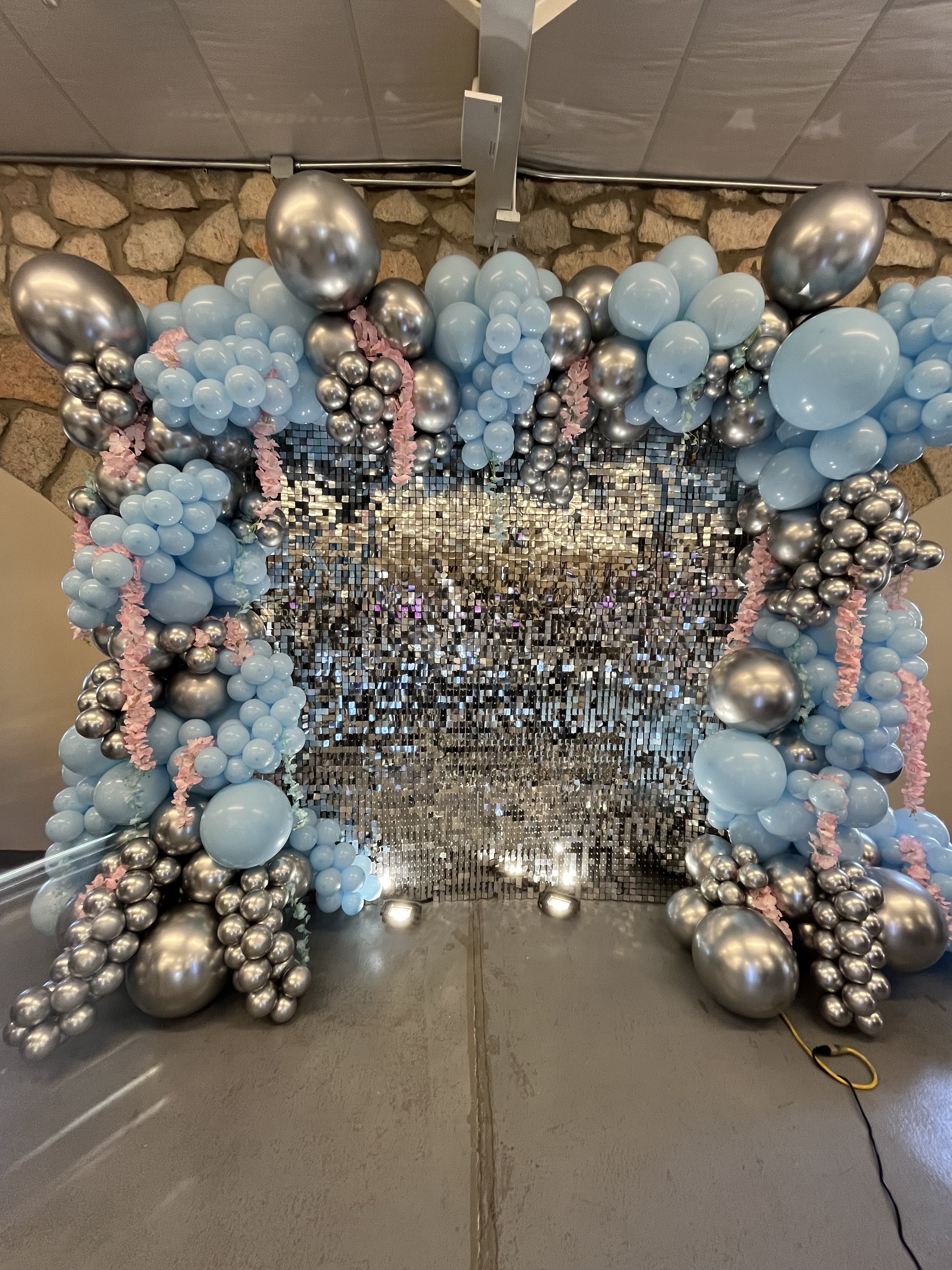 Decorative balloon and sequin backdrop for an event or celebration with blue, silver, and pink balloons arranged in clusters, with a sequined wall behind it.