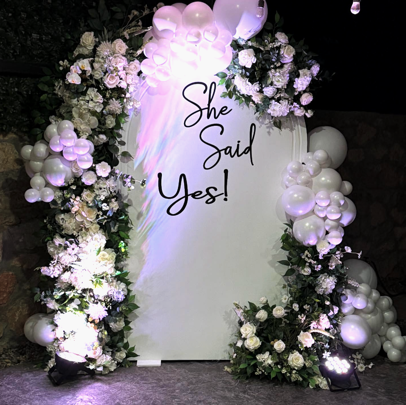 Arco decorativo de globos blancos y flores blancas y verdes, con cartel que dice 'She Said Yes!' en letras negras, con luces de escenario en el suelo.