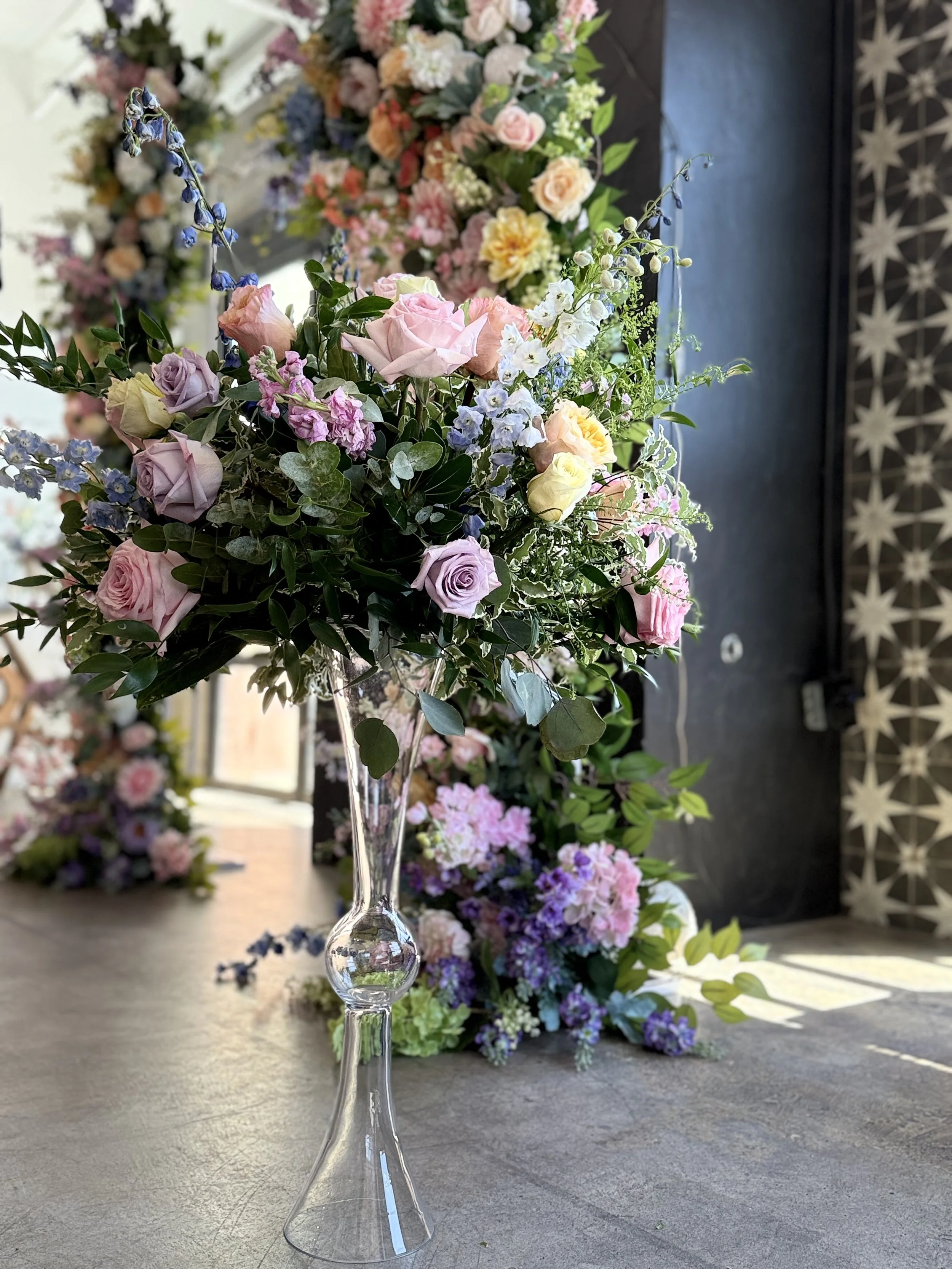 A floral arrangement with pink, purple, white, and yellow roses, and greenery in a tall, clear glass vase on a gray surface, with additional flowers and patterned wall in the background.