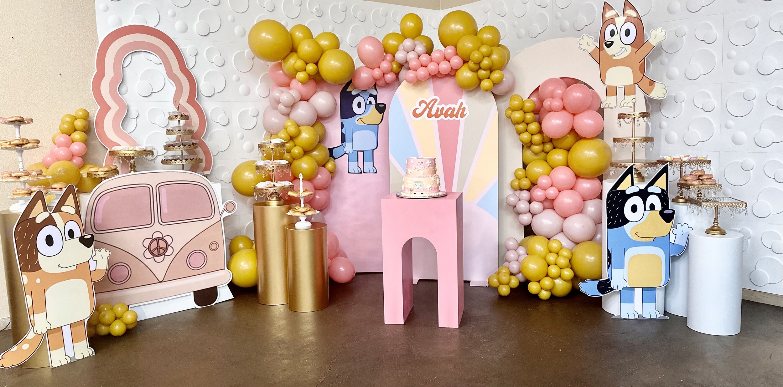 A colorful party setup with cartoon dog characters, balloons, and a pink cake. The backdrop features cartoon dog faces and a pastel rainbow. There are tiered trays of cookies on gold stands and a pink table with a cluster of pink and yellow balloons,